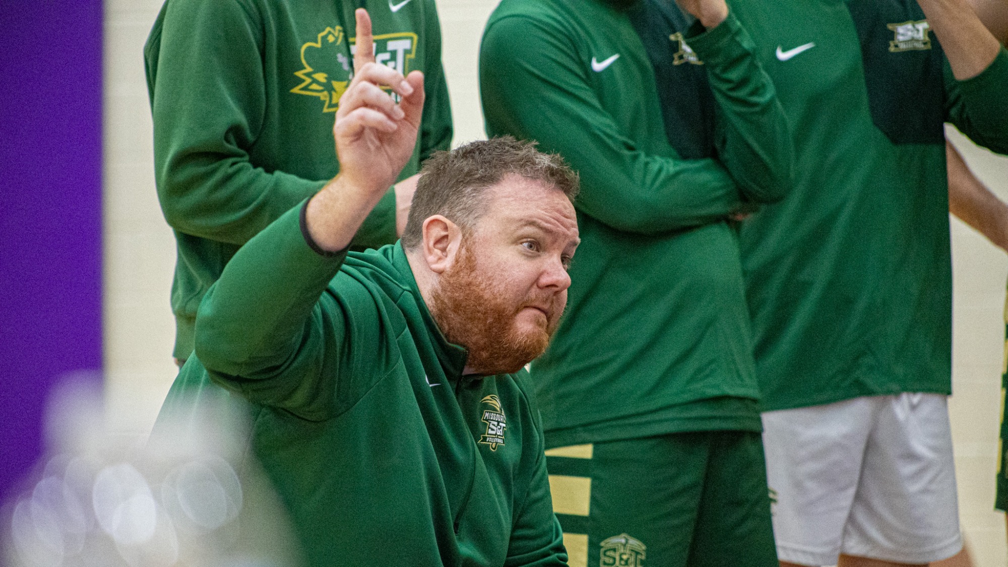 Missouri S&T Men's Volleyball Head Coach Ryan Thompson kneeling during a team huddle during a match, addressing players and pointing at something in the distance. He is wearing a green Missouri S&T pullover and is surrounded by S&T student-athletes. 