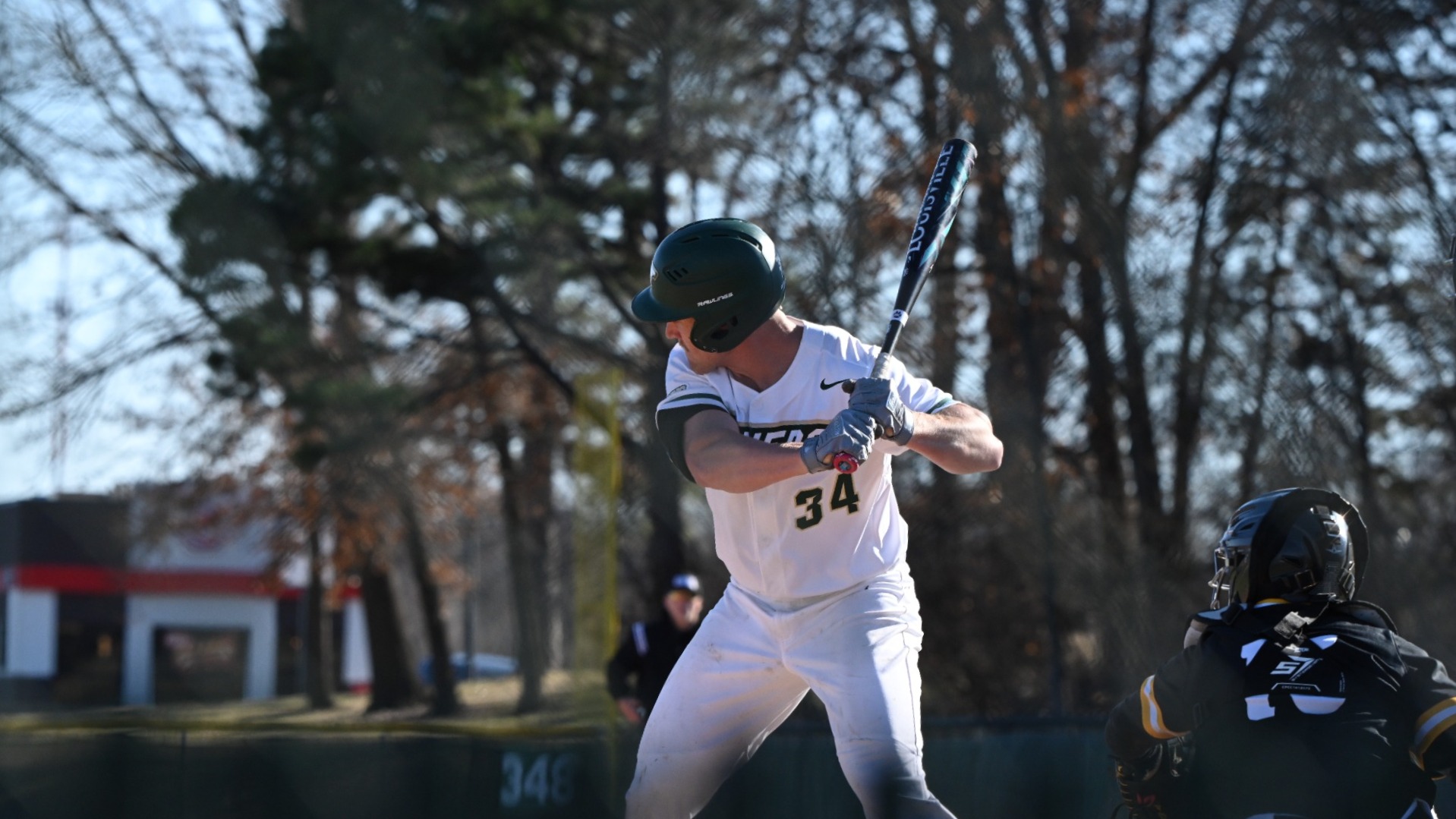 Will Beckham photographed in a right-handed batting stance at home plate, wearing a white Missouri S&T uniform with number 34 and a green helmet, holding the bat high over his shoulder as a catcher in black gear sets up behind him on a sunny day with trees in the background.