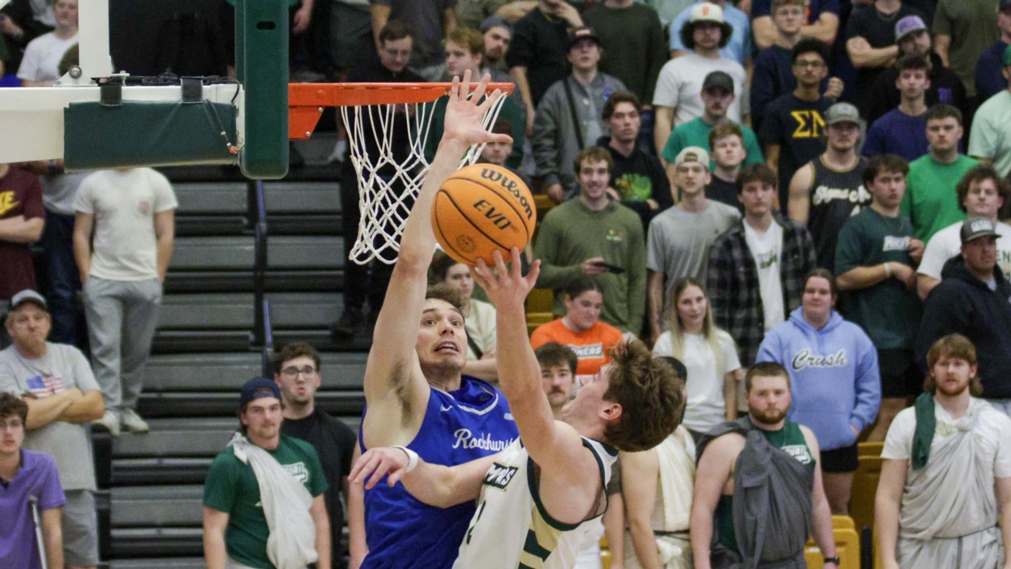 Missouri S&T's Alex Benassi, wearing a white and green jersey, leaps toward the basket for a layup. He is closely contested by a Rockhurst player in a blue jersey who has his arm raised high to block the shot. The orange basketball is suspended in the air between them, just below the rim. In the background, a dense crowd of students in the bleachers watches the play with expressions of intense focus and excitement.