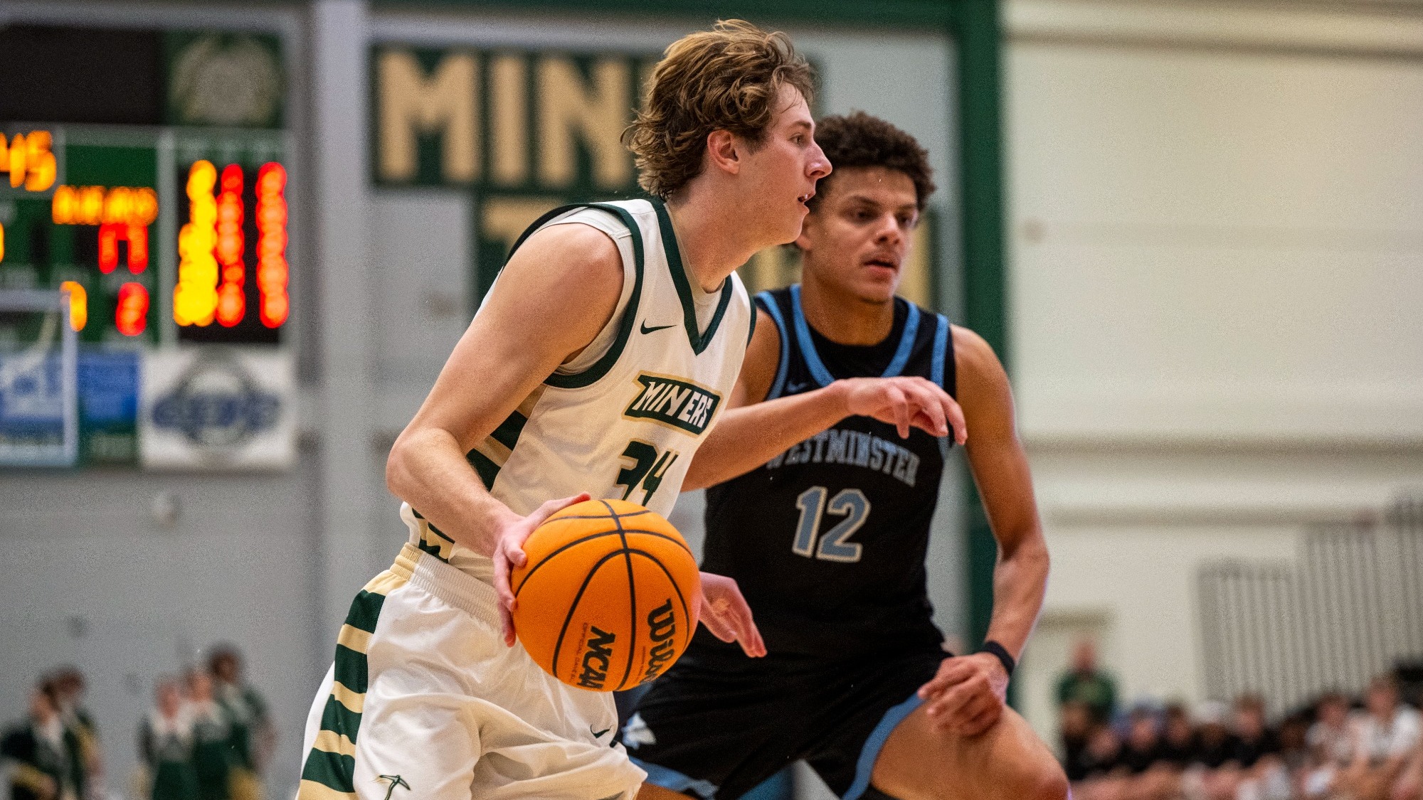 Andrew Young, photographed in a white and green Miner basketball uniform, dribbles the ball with his right hand while driving toward the lane as a defender in a black Westminster jersey closely guards him.