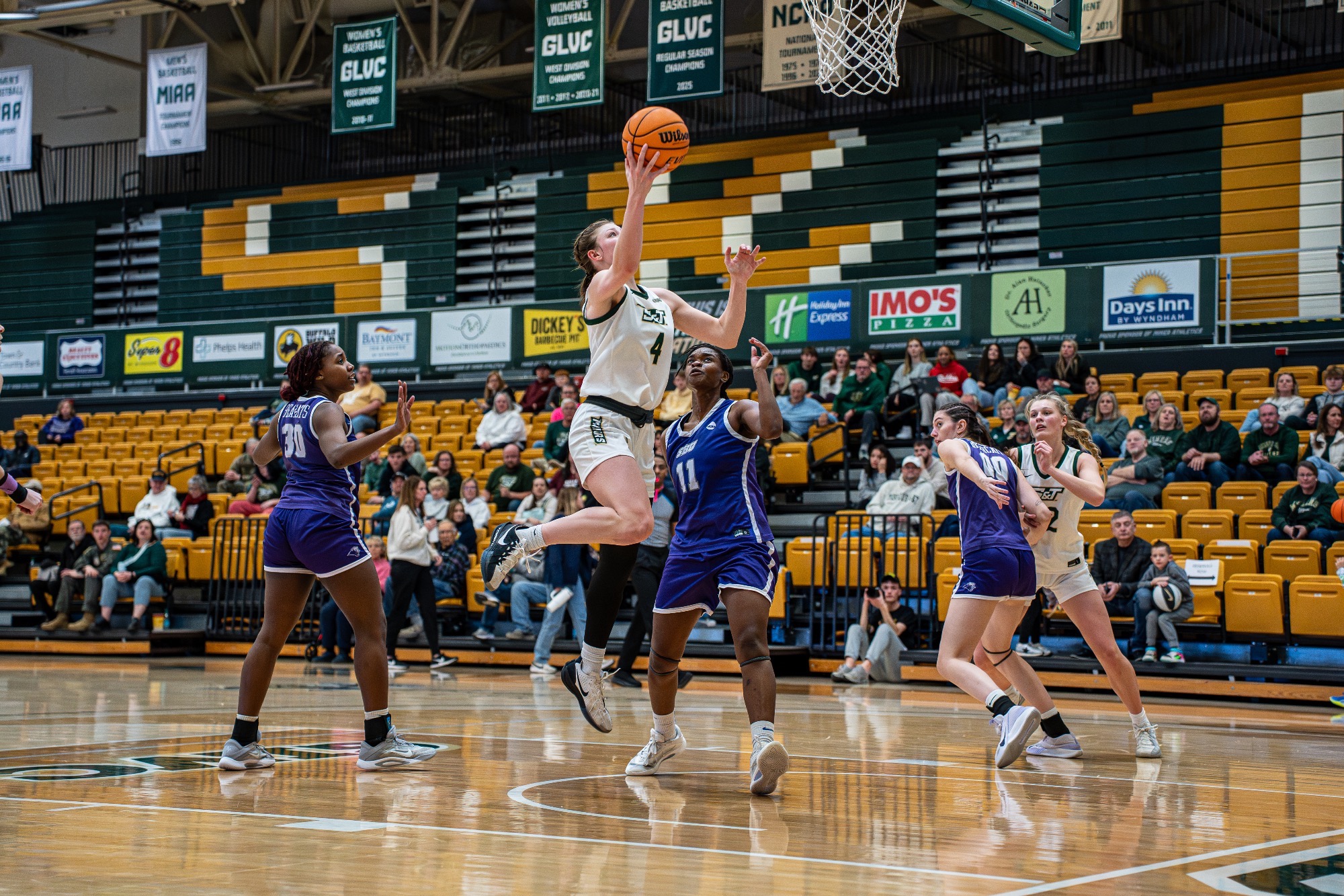 Missouri S&T women's basketball player Norah Gum in a white S&T jersey going up for a layup in Gibson Arena with a SBU player in purple guarding her.