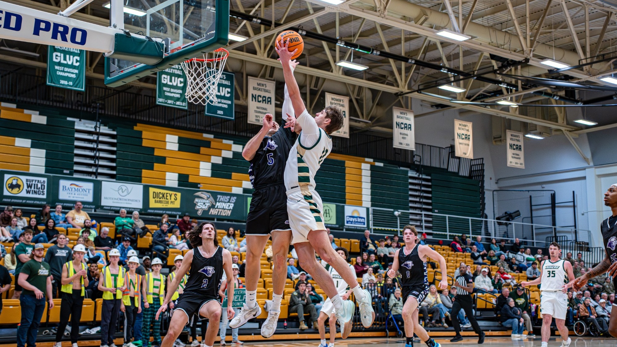 Oliver Kokal of the Missouri S&T Miners is photographed midair driving toward the basket for a right-handed layup, extending above a defender in a black jersey who contests the shot. The action takes place inside Gibson Arena, with green and gold seating, GLVC and NCAA banners, and fans watching from the stands in the background.