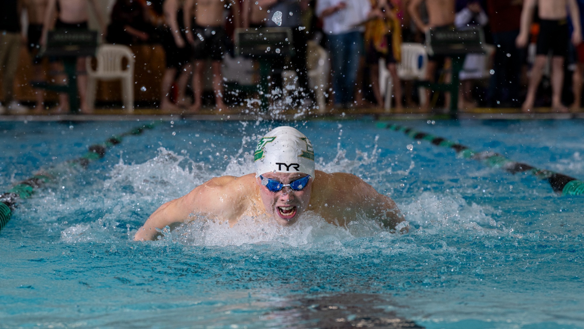 John Cottrell photographed mid-stroke in the butterfly during an indoor swim meet, wearing a white Missouri S&T swim cap and blue goggles as water splashes around him, with teammates and spectators blurred in the background.