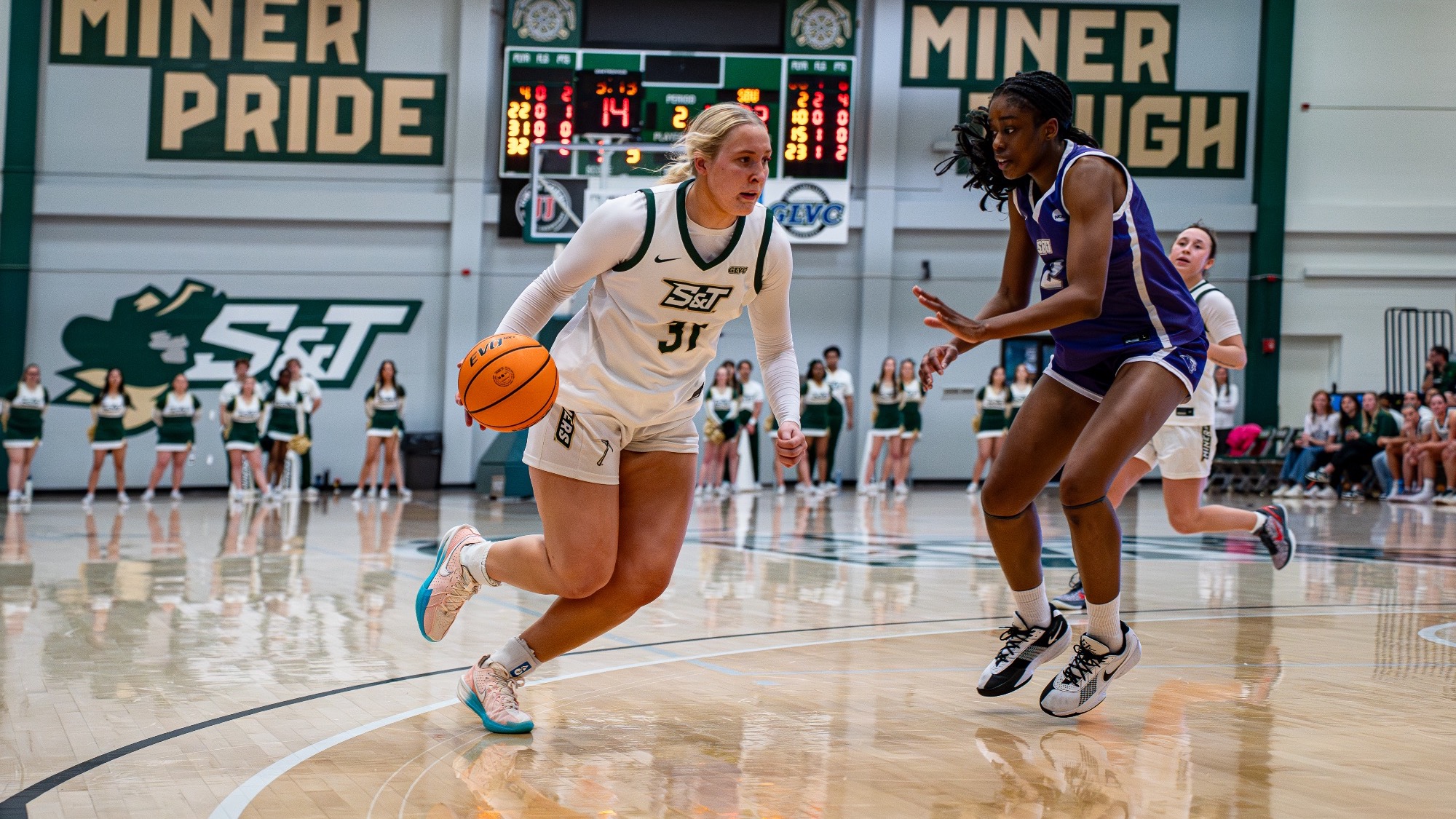 Missouri S&T forward Anna Gilbertson in a white S&T jersey dribbling the ball to the paint with an SBU defender on her in a purple Jersey in Gibson Arena.