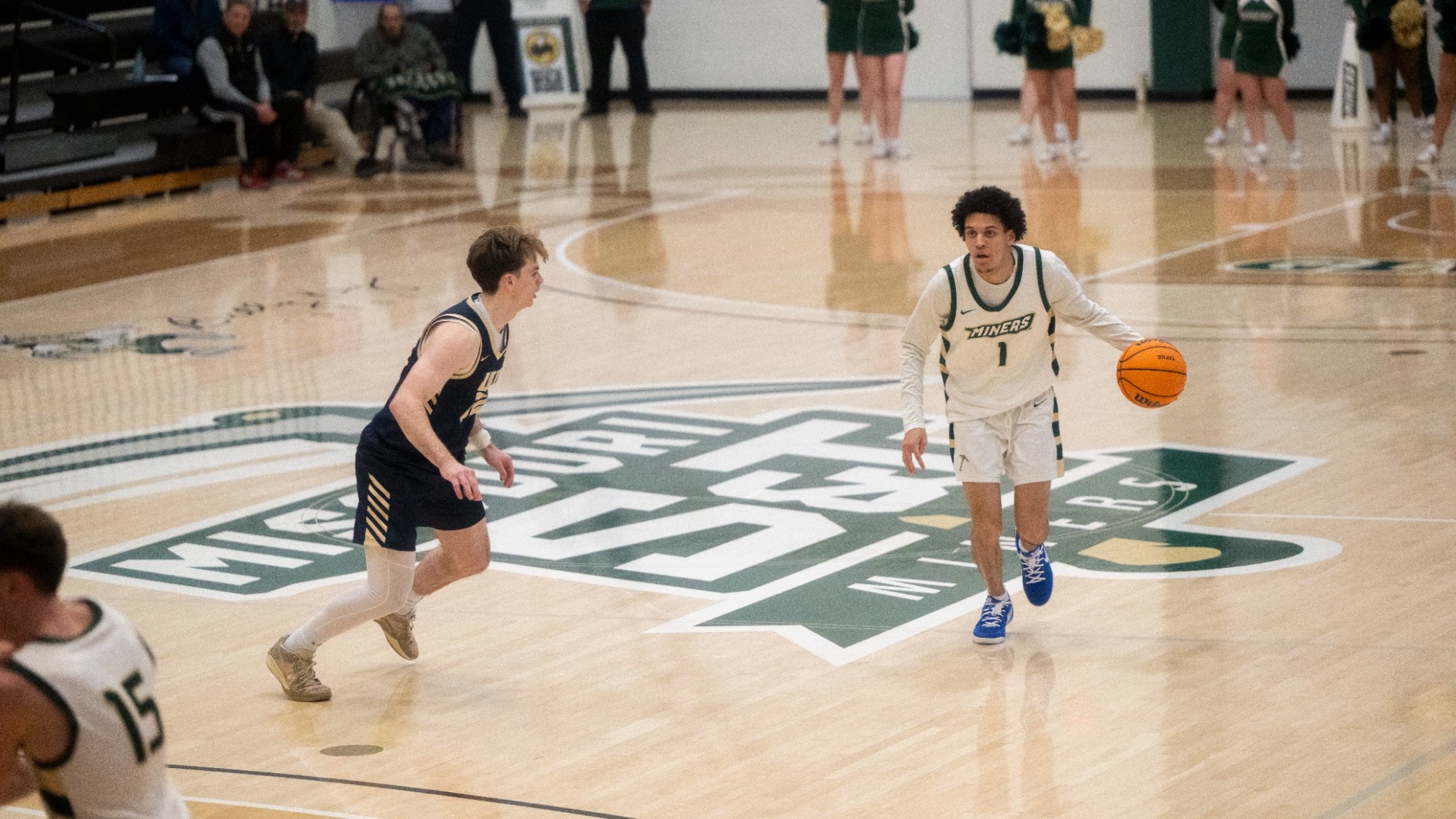 Blaise Beauchamp is photographed dribbling the basketball with his right hand near midcourt during a home game. He wears a white Missouri S&T Miners uniform with green trim and blue shoes, facing a defender in a navy uniform who is crouched in front of him. The large Miners logo is visible at center court, with cheerleaders and spectators standing along the baseline in the background.
