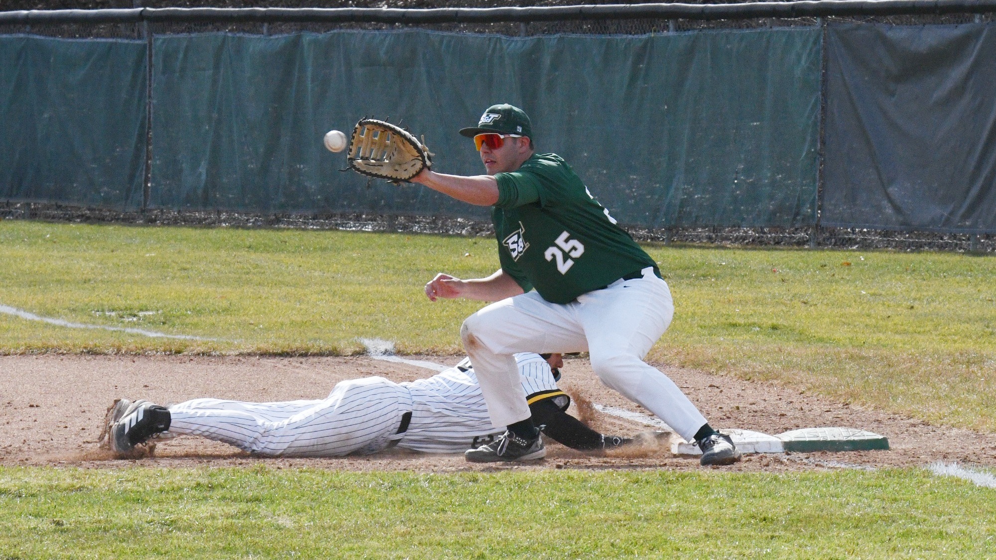 Nick Hooper, photographed in a green Missouri S&T baseball jersey and white pants, stretches toward an incoming throw at first base with his glove extended as a runner in a white pinstriped uniform slides headfirst back to the bag, kicking up dirt during a close play.