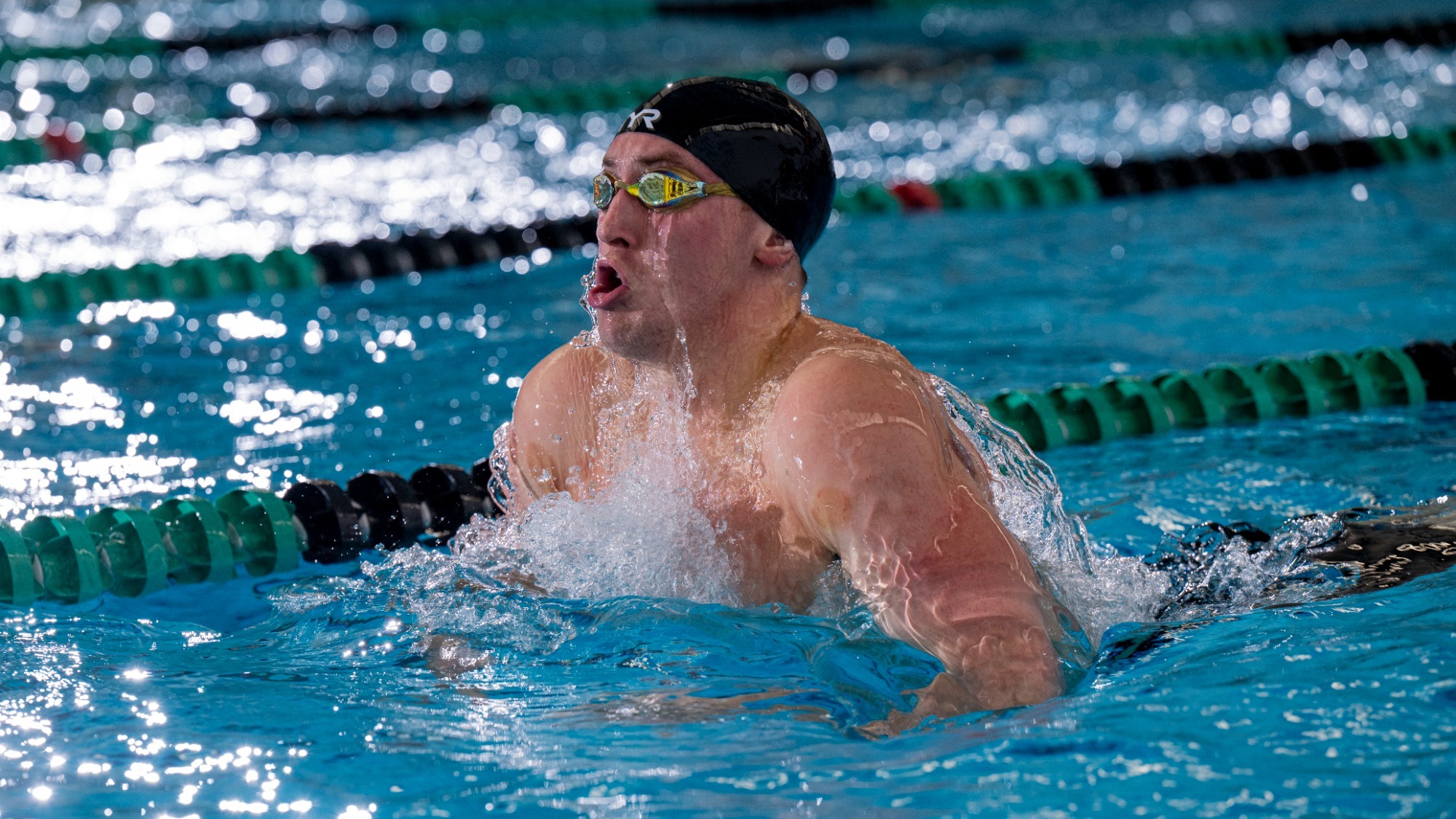 Connor Bichsel is photographed swimming the breaststroke during a meet, wearing a black swim cap and mirrored goggles as he rises out of the water for a breath. Water splashes around his shoulders and chest as he powers forward between green and black lane lines in an indoor pool.