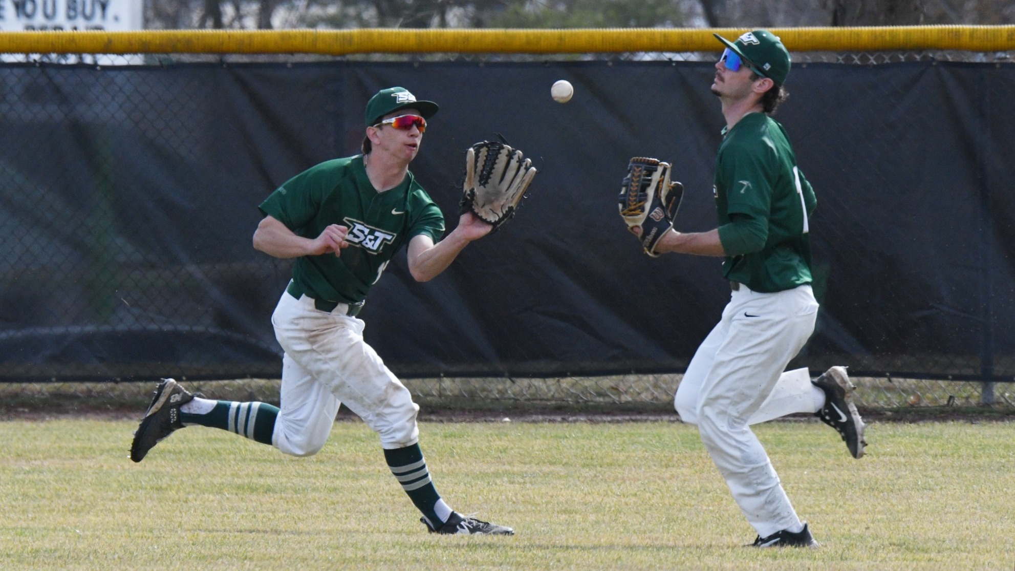 Garrett Meyer (right) and Drew Baugus (left) are photographed converging in the outfield during a baseball game, both wearing green Missouri S&T jerseys and white pants as they track a fly ball descending between them. Baugus runs forward with his glove extended while Meyer moves in from the right with his glove raised, the outfield fence visible in the background.