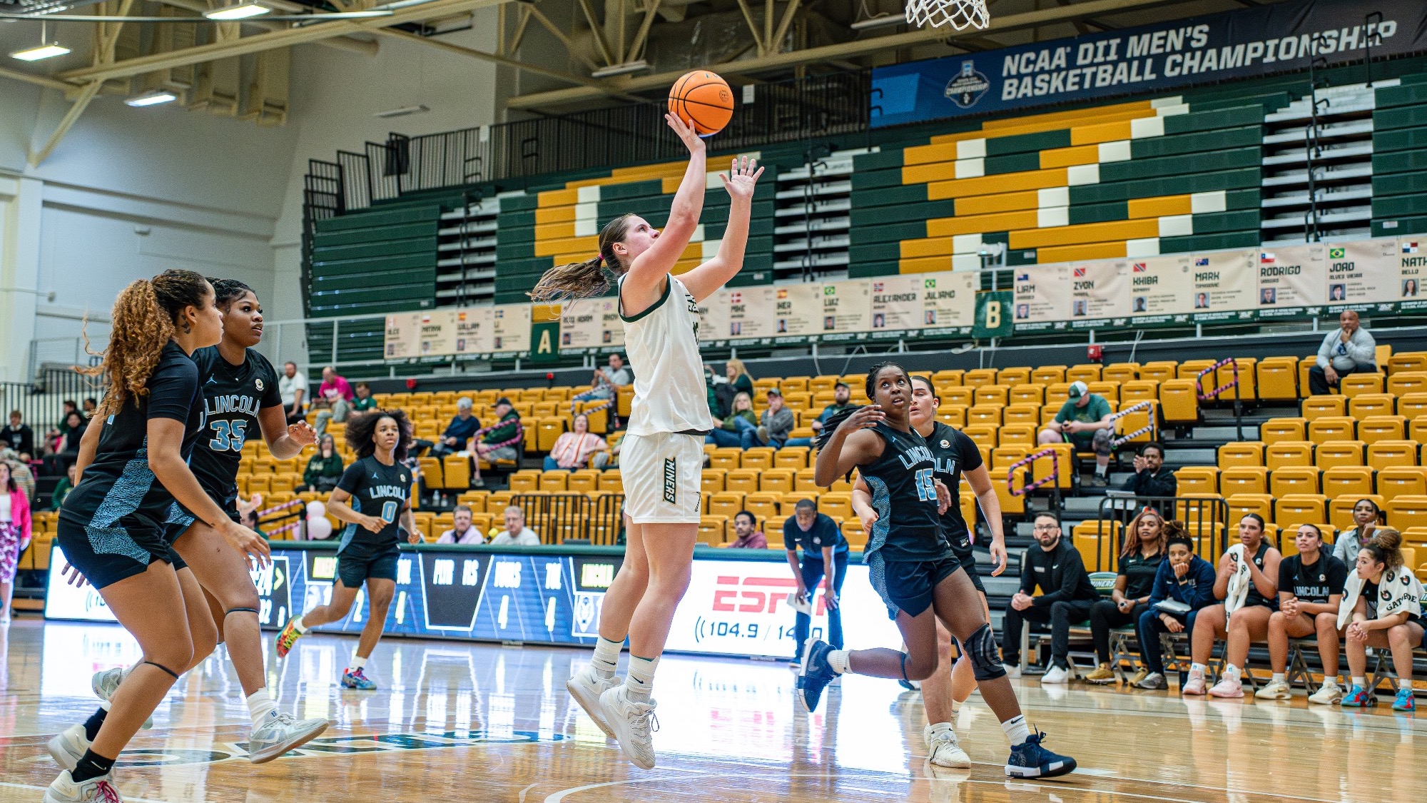 Missouri S&T forward Megan Aulbert, wearing a white Miners jersey, elevates for a right-handed layup inside Gibson Arena while a Lincoln University defender in a blue uniform contests from her right, with teammates and fans visible in the background.