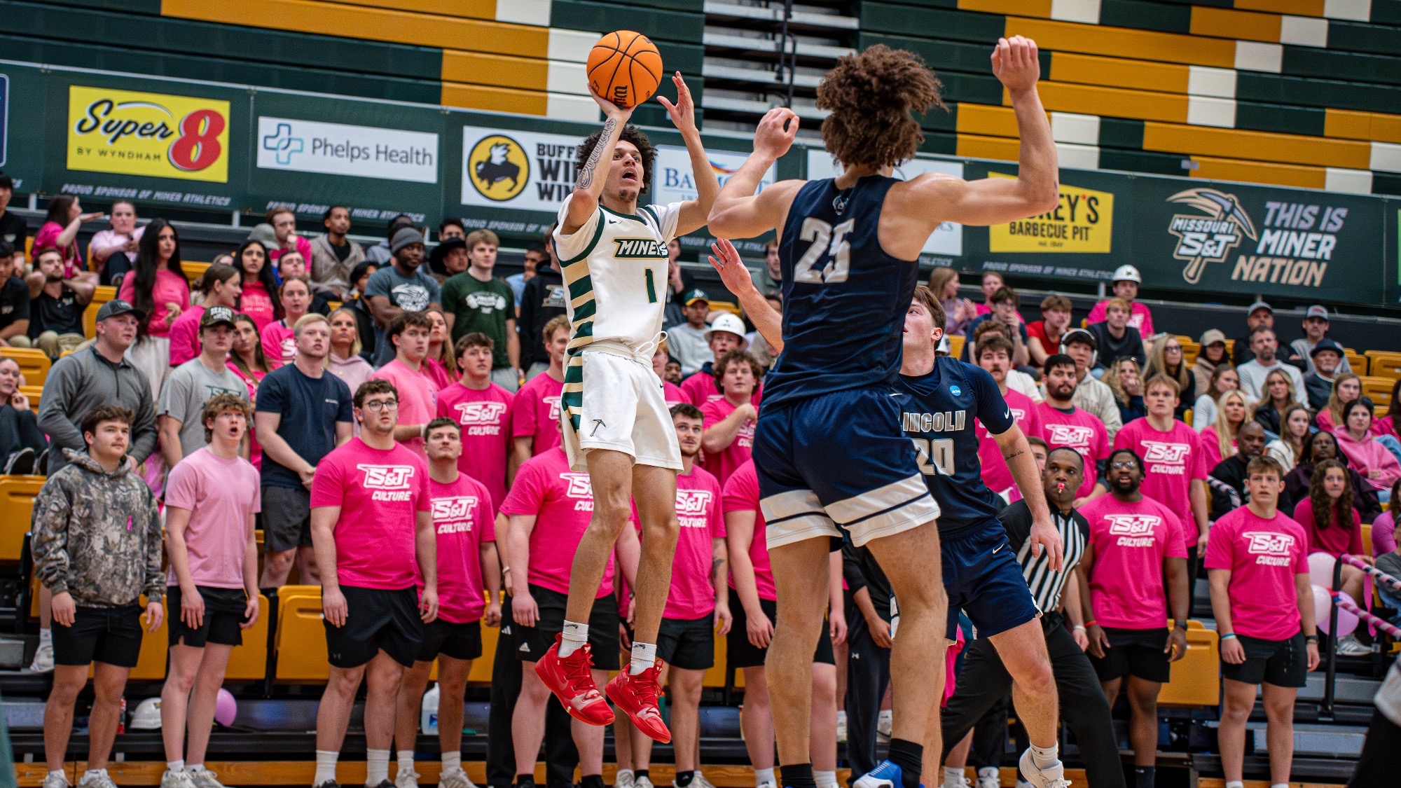 Missouri S&T guard Blaise Beauchamp, wearing a white Miners jersey, elevates for a pull-up jumper in the lane while a Lincoln defender in a blue uniform contests with a hand up, as a packed student section in pink shirts watches from behind the basket.