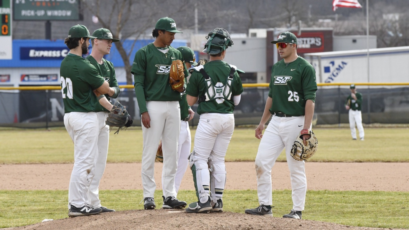 Six Missouri S&T baseball players in green and white uniforms hold a meeting on the pitcher's mound during a game. The background shows an electronic scoreboard, outfield fences with local advertisements, and an American flag under a clear sky.