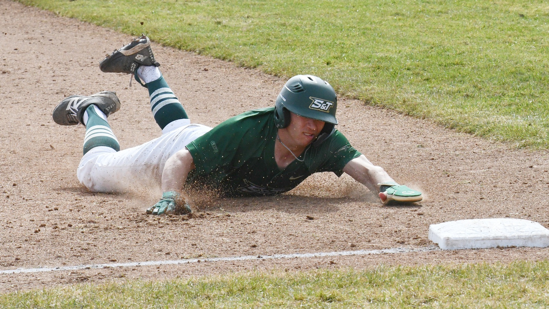 A Missouri S&T Miners baseball player, Drew Baugus, mid-dive as he slides headfirst into a base on a dirt infield. He is wearing a dark green jersey with 
