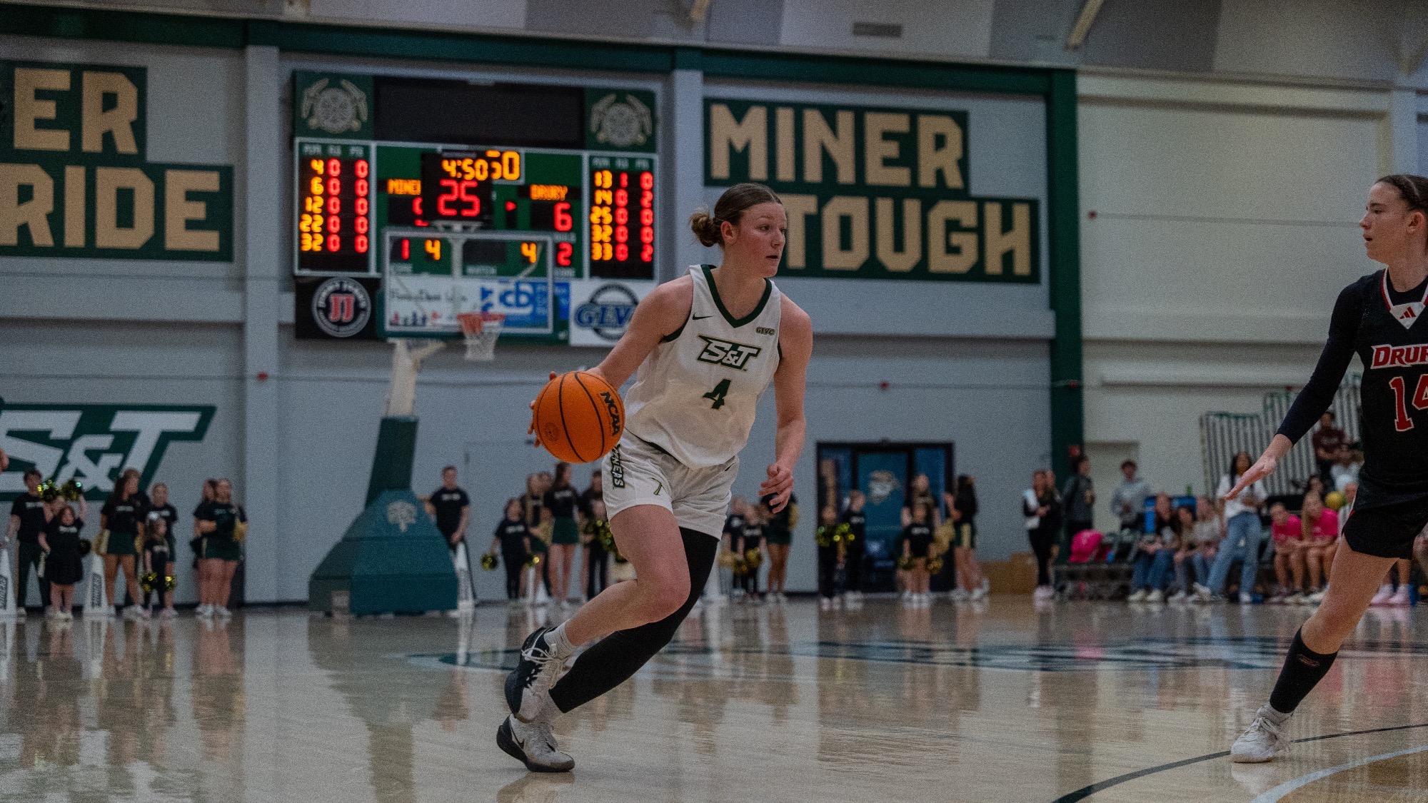 Norah Gum of the Missouri S&T Miners dribbles the basketball up the court during a home game, wearing a white No. 4 jersey, as a defender approaches and teammates and fans watch from the background beneath a “Miner Tough” sign and scoreboard.