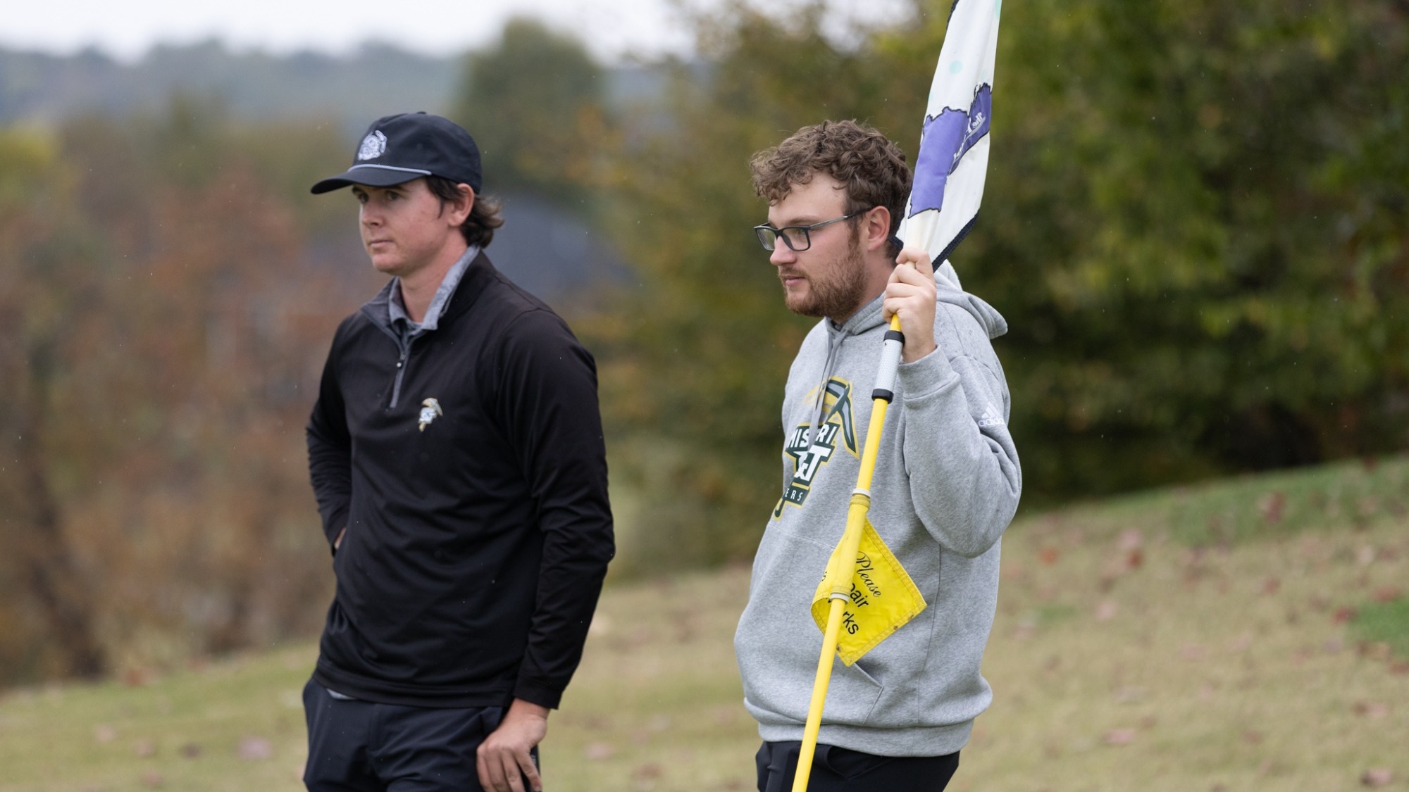Cole Komyati, photographed on the left, and assistant coach Jack Coleman, photographed on the right, stand on a golf course during competition. Komyati wears a black cap and black pullover while holding a golf club and looking ahead. Coleman wears glasses and a gray Missouri S&T hoodie while holding a yellow flagstick, with trees and rolling terrain visible in the background.