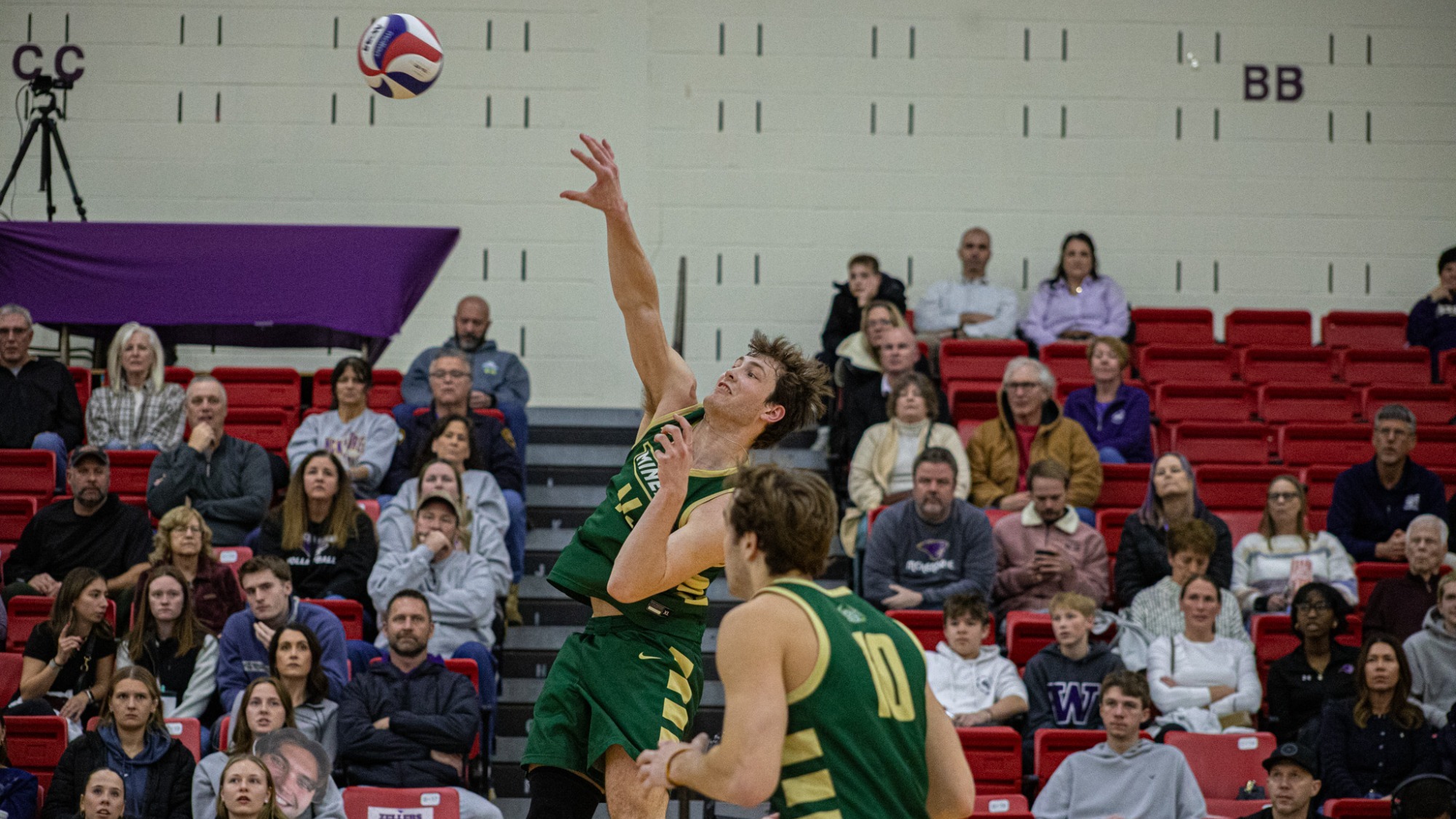 Missouri S&T men's volleyball athlete Aaron Sallade is photographed wearing a green uniform and jumping and attacking a volleyball during a match at McKendree
