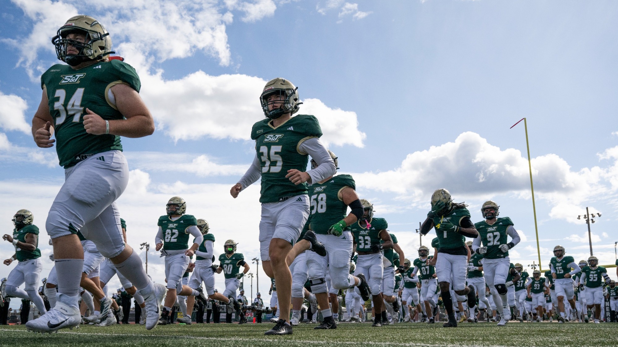 Missouri S&T Miners football players are **photographed** running onto the field together during pregame, wearing green jerseys and white pants, with helmets on as they jog across the turf under a partly cloudy sky. A goalpost is visible in the background, emphasizing the game-day setting and team unity as the Miners take the field.