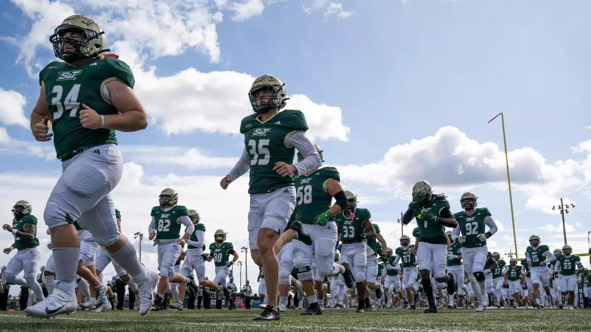 Missouri S&T Miners football players are photographed running onto the field together during pregame, wearing green jerseys and white pants, with helmets on as they jog across the turf under a partly cloudy sky. A goalpost is visible in the background, emphasizing the game-day setting and team unity as the Miners take the field.