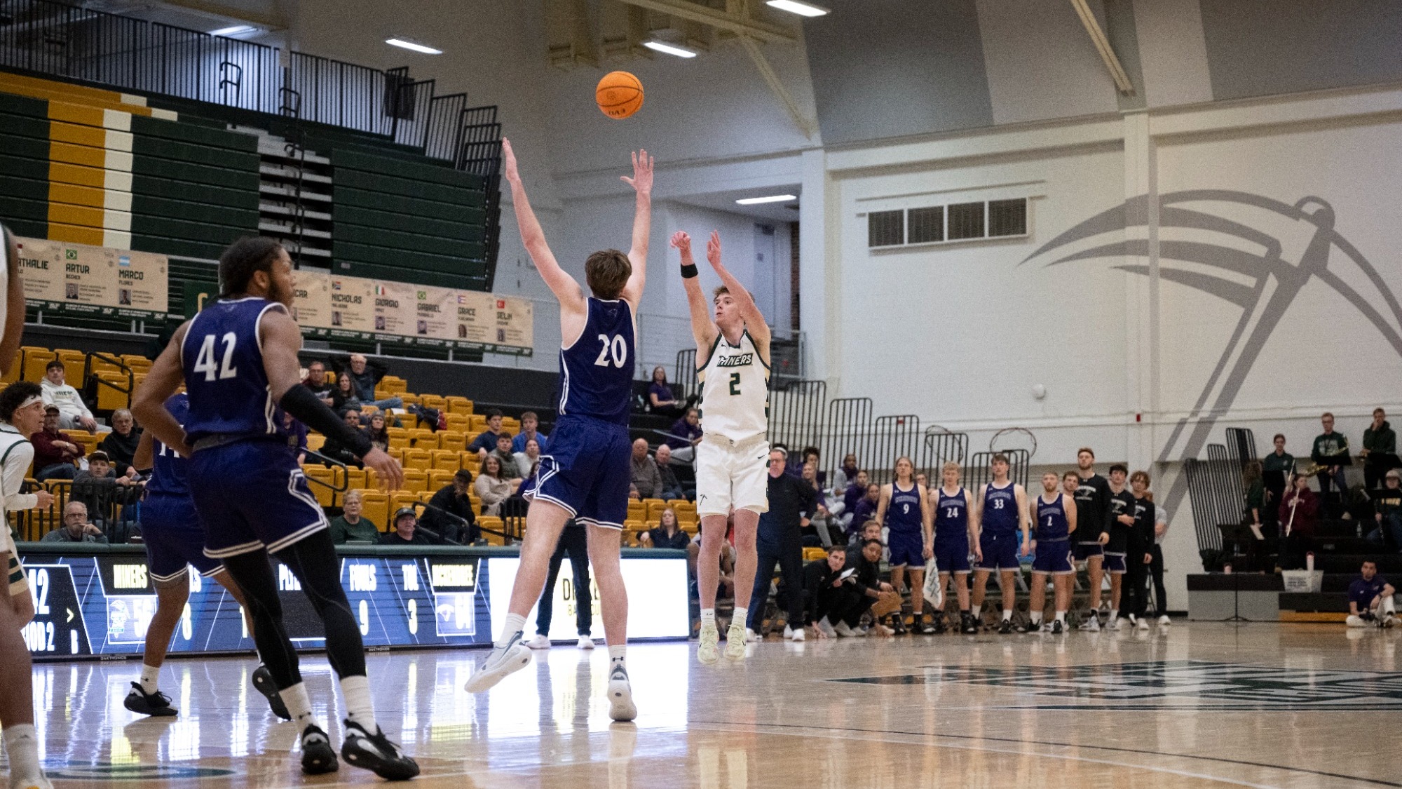 Missouri S&T men's basketball player Alex Benassi wears a white jersey and matching pants. He is pulling up for a three-point jump shot while a McKendree defender jumps to try and block the shot with his hand outstretched during a game in Gibson Arena