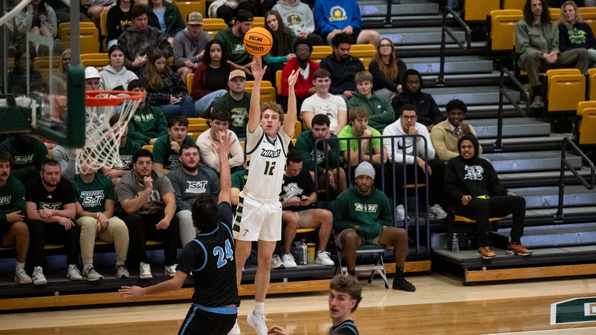 Missouri S&T men's basketball athlete Aidan Burns is shooting a three-pointer during a game in Gibson Arena with the goal visible in the foreground, fans visible behind him, and an opposing defender closing the gap to try to block the shot