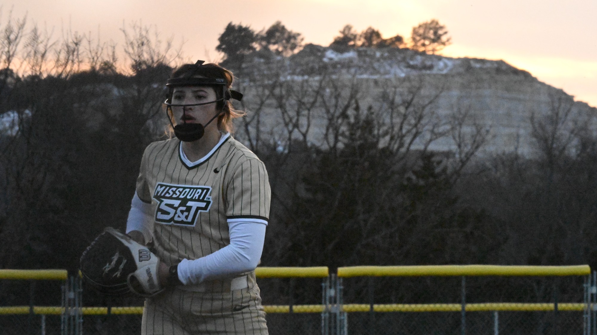 Missouri S&T pitcher AJ Agers prepares to deliver a pitch, wearing a gold Miners jersey in the circle with a mountainous backdrop beyond the outfield fence.
