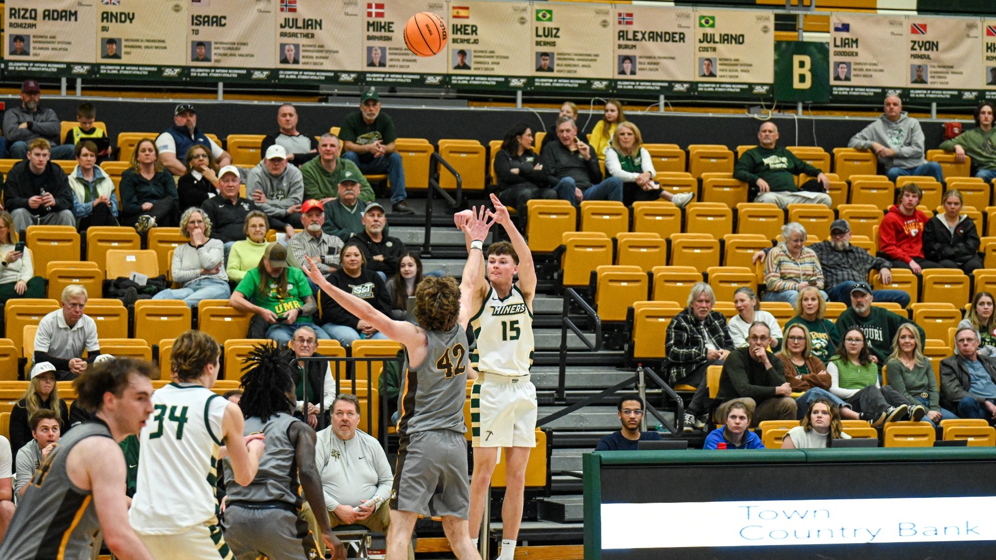 Missouri S&T men’s basketball player Garrett Hines rises for a three-point shot in a white Miners jersey as a Quincy defender in a gray jersey contests the attempt during a game inside Gibson Arena.