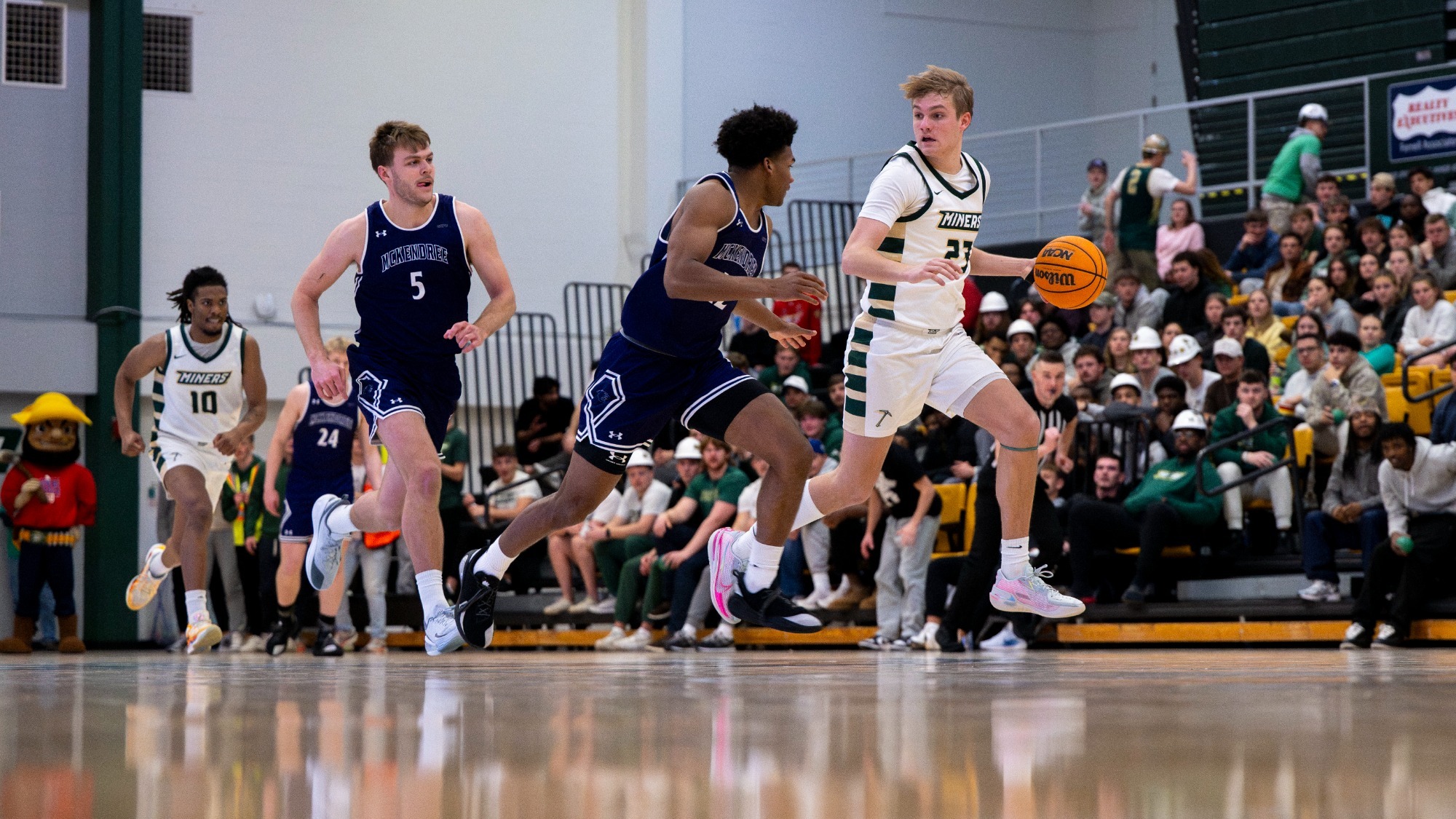 Missouri S&T men's basketball athlete Adam Rickman dribbling the basketball with his left hand as he dribbles away from two McKendree defenders with fans and a teammate visible in the background during a game in Gibson Arena
