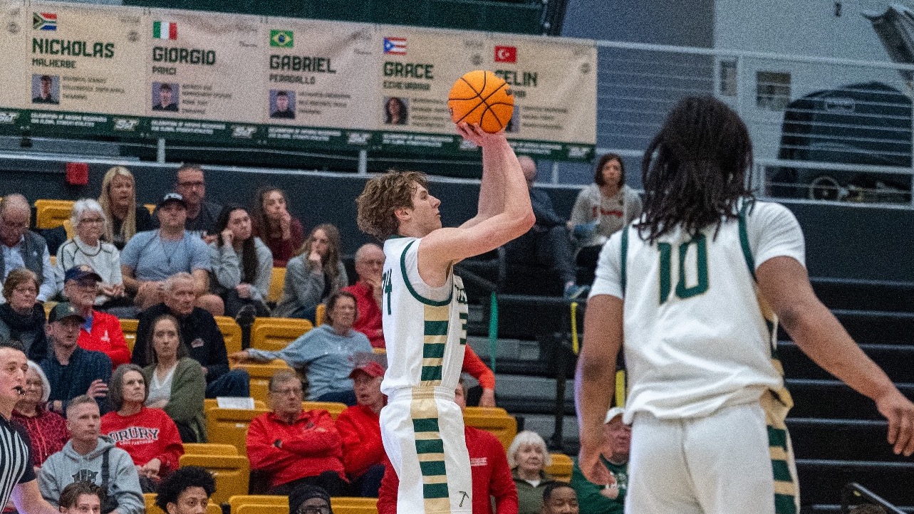 Missouri S&T Men’s Basketball player Andrew Young wearing green and white jersey while shooting a three-poiinger during a home game in Gibson Arena. Fans are visible in the background.
