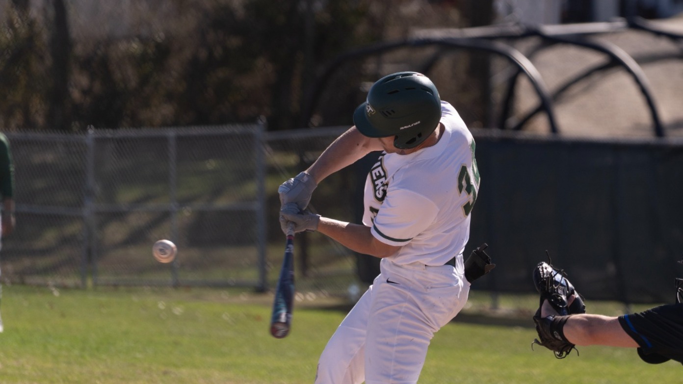 Will Beckham is photographed swinging the bat and about to make contact with the baseball during a Missouri S&T game, his body turning through the swing as the opposing catcher reaches forward from behind home plate to field the pitch.