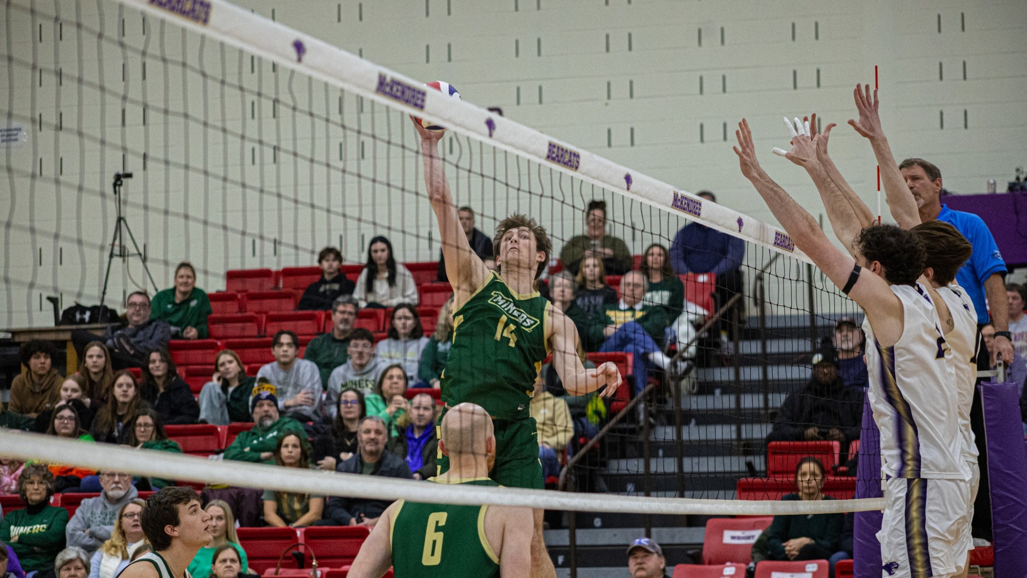 Aaron Sallade of Missouri S&T is photographed leaping at the net to attack the ball with his right hand during a men’s volleyball match, as two opposing blockers in white jerseys jump with their hands raised to defend. Two Miners teammates watch from the court in the foreground while spectators sit in red bleachers behind the play.