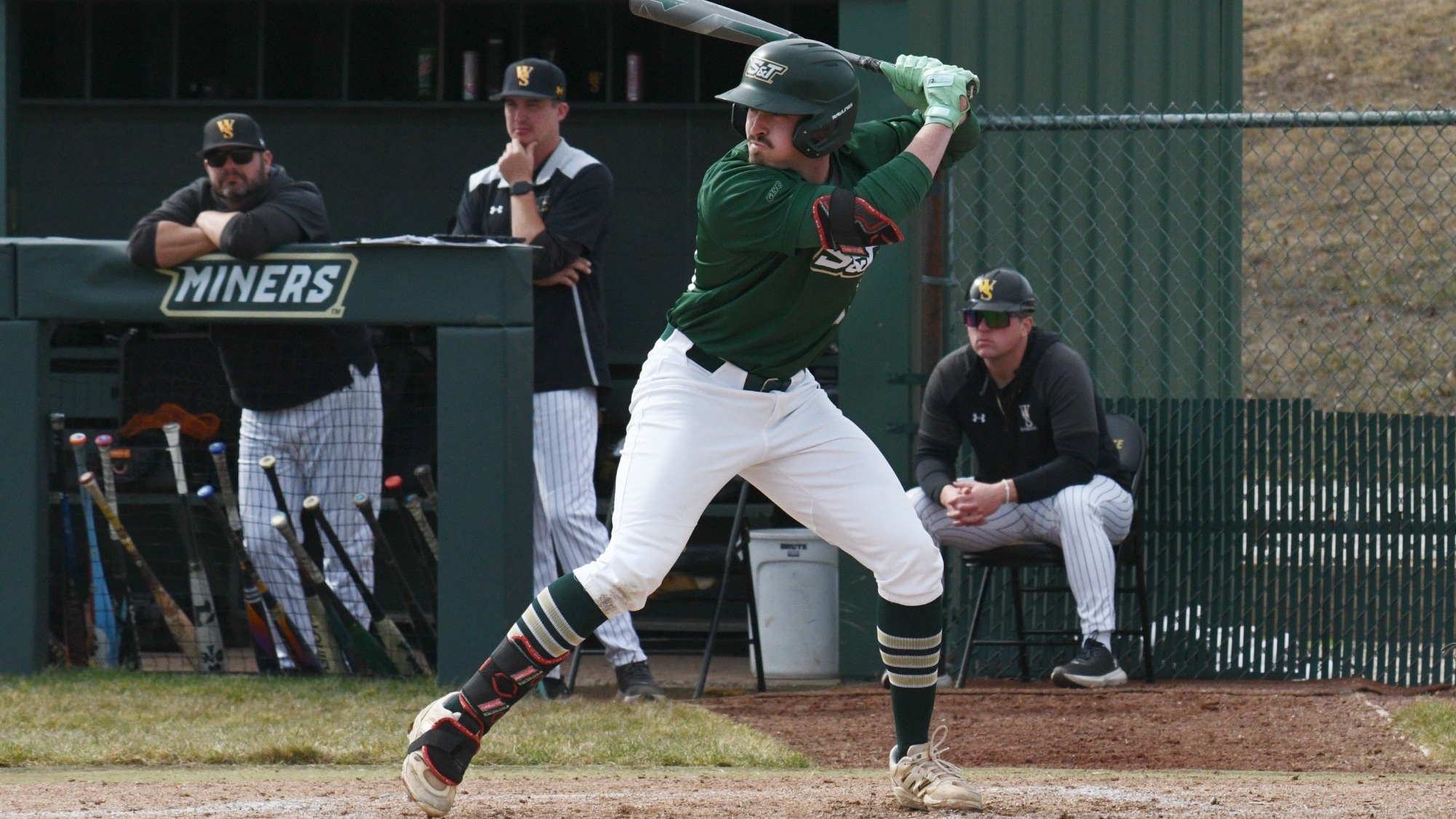 Missouri S&T baseball player Caleb Horsey stands in the batter’s box mid-swing during a game, photographed wearing a green Miners jersey, white pants, and a batting helmet. Horsey holds the bat high as he begins his swing, with the Missouri S&T dugout, teammates, and bat rack visible behind him along the fence line.