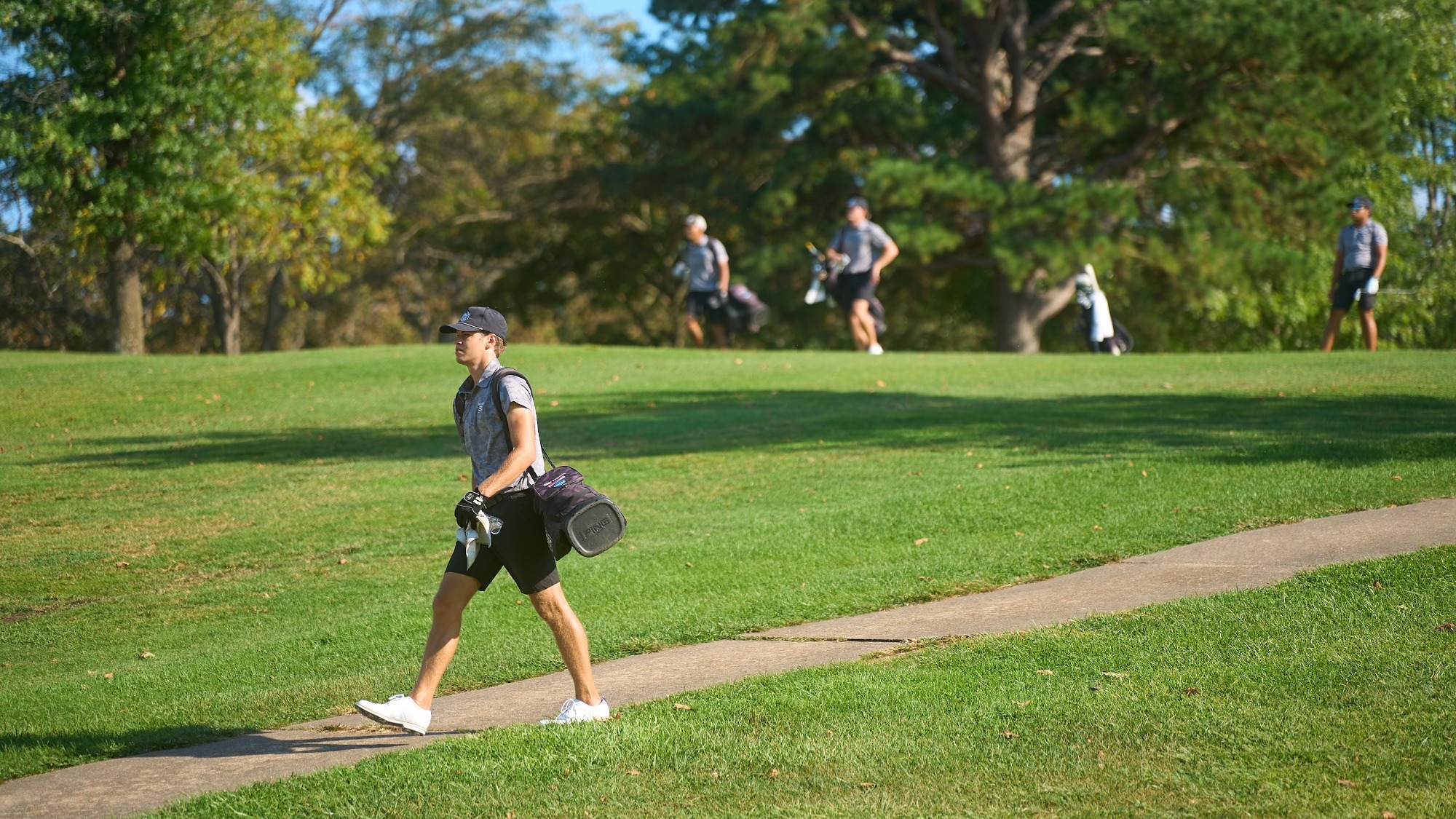 Joey Friedel, photographed walking along a paved path on a golf course while carrying his golf bag over his shoulder. He wears a gray polo, black shorts, a black cap, and white golf shoes. In the background, three additional golfers with bags walk across the fairway near a tree line on a sunny day.
