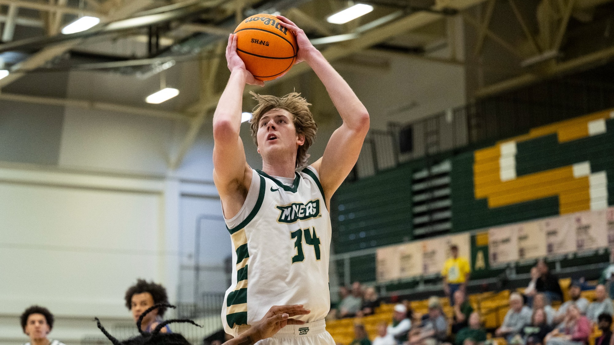 Andrew Young, a basketball player for the Missouri S&T Miners, is shown mid-air in a white home jersey with green and gold accents and the number 34.  He is focused on the rim, holding a Wilson basketball high above his head with both hands in a shooting motion. A defender with long dark hair and a tattooed arm is seen from behind in the foreground, reaching up to contest the shot. In the background, teammate #1 watches from the court, and the gymnasium stands are filled with fans in green and gold.