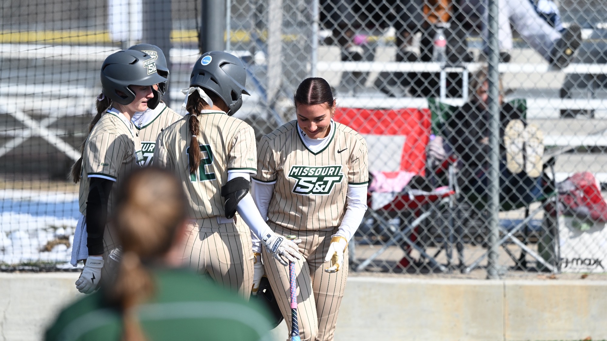 Keira Jones Missouri S&T softball player in gold pin stripes jersey celebrating with her teammates at home plate after hitting a homer during a softball game.