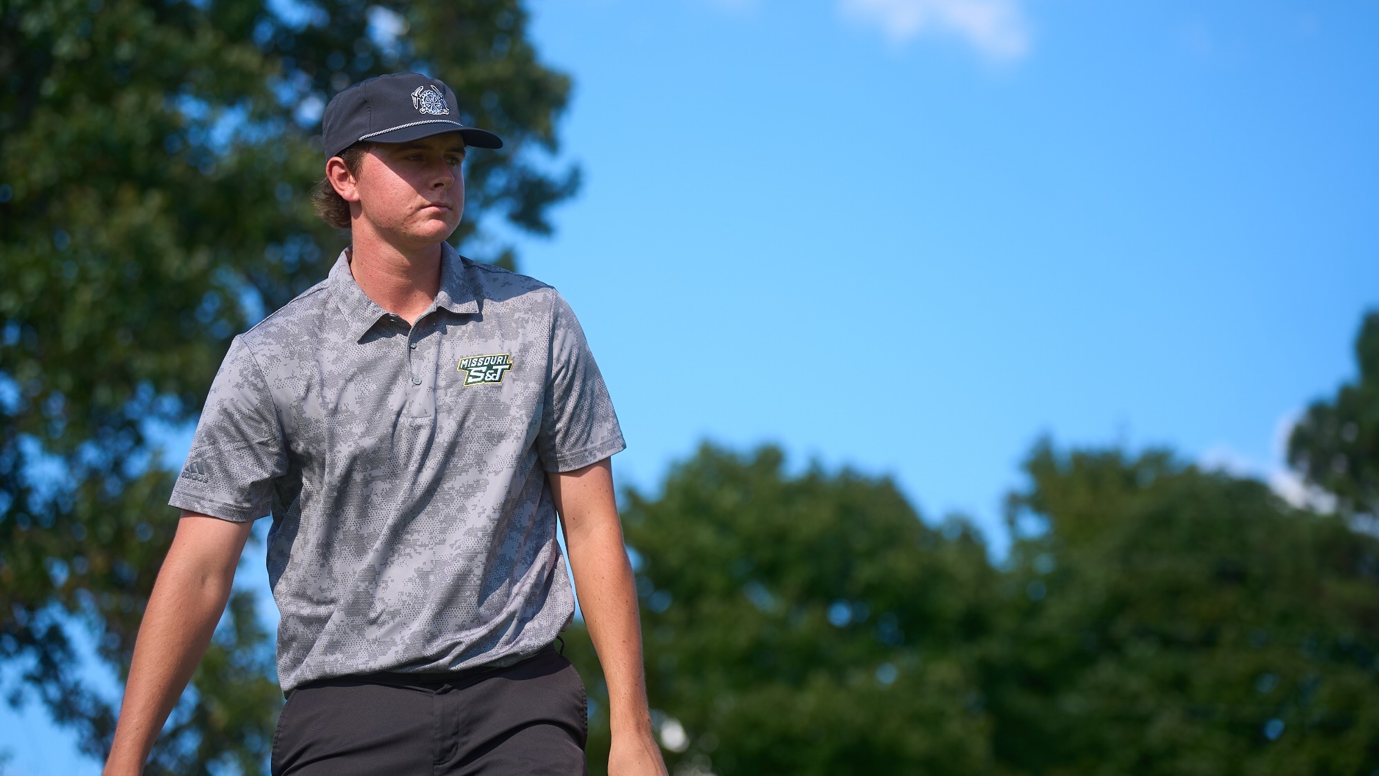 A medium shot of golfer Cole Komyati of Missouri S&T walking outdoors on a sunny day. He is wearing a grey patterned polo shirt with the Missouri S&T logo and a dark baseball cap. He is looking off to the side with a focused expression against a background of green trees and a blue sky.