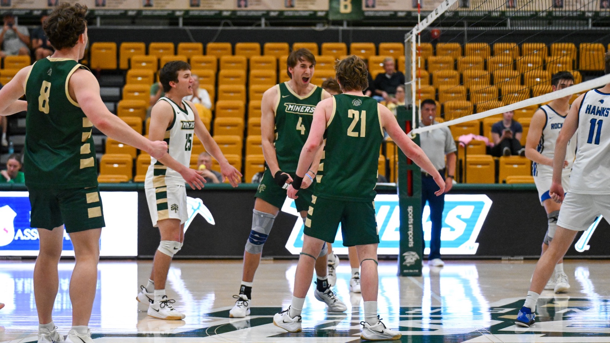Indoor men’s volleyball match in a gymnasium. Four players in dark green “Miners” uniforms celebrate near the net after a point; player #4, Tyler Johnwick, stands at center shouting with intensity, knees bent and fists clenched. Teammates #8, #21, and #25 face him with arms out, appearing energized. Opponents in white uniforms stand on the opposite side of the net. Yellow arena seats and banners line the background. 