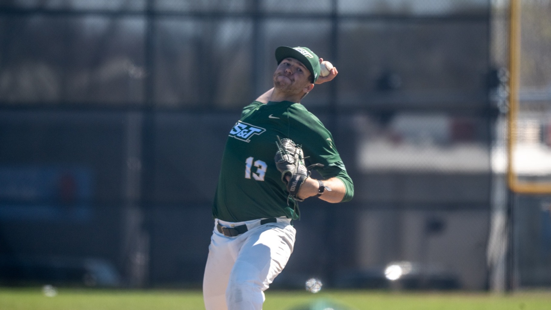 Dylan Bates, a Missouri S&T baseball pitcher wearing a green jersey with number 13 and a green cap, is photographed mid-throw on the mound, his arm extended back with the ball in hand as he begins his pitching motion. He wears white pants and a glove on his opposite hand, with the outfield fence and a sunlit field softly blurred in the background.