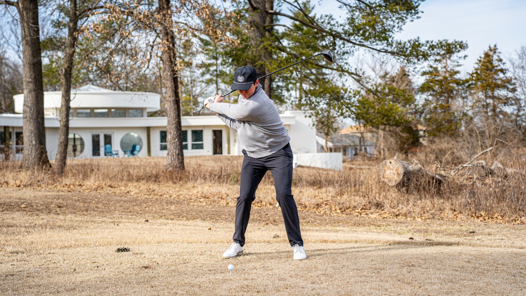 Cole Komyati is photographed on a golf tee box mid-backswing, preparing to drive the ball while wearing a black cap, gray long-sleeve top, dark pants, and white golf shoes. A golf ball sits on a tee in front of him on dry grass, with trees and a clubhouse visible in the background on a clear day.