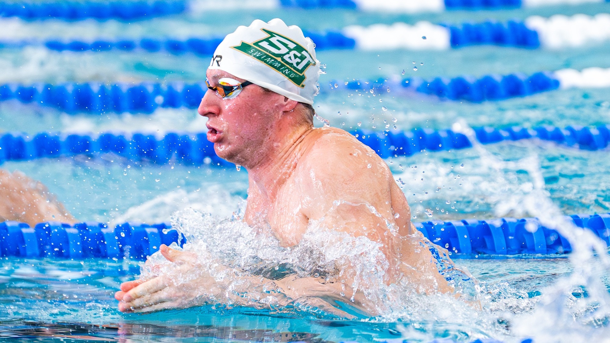 Missouri S&T Swimmer Jordan Christensen coming out of the water while doing the breaststroke in the pool at the national meet. He is wearing a white swim cap with the Missouri S&T logo on it. He is also wearing swim goggles
