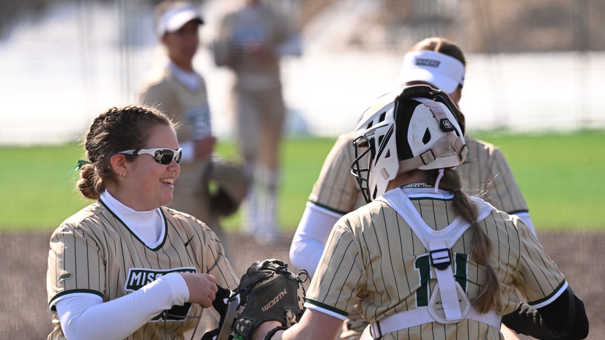 Missouri S&T softball pitcher Riley Wilkinson, wearing a gold pinstripe Miners jersey and sunglasses, smiles while talking with catcher Trista Hunt, who stands with her back to the camera in full catcher’s gear, during a moment on the field.