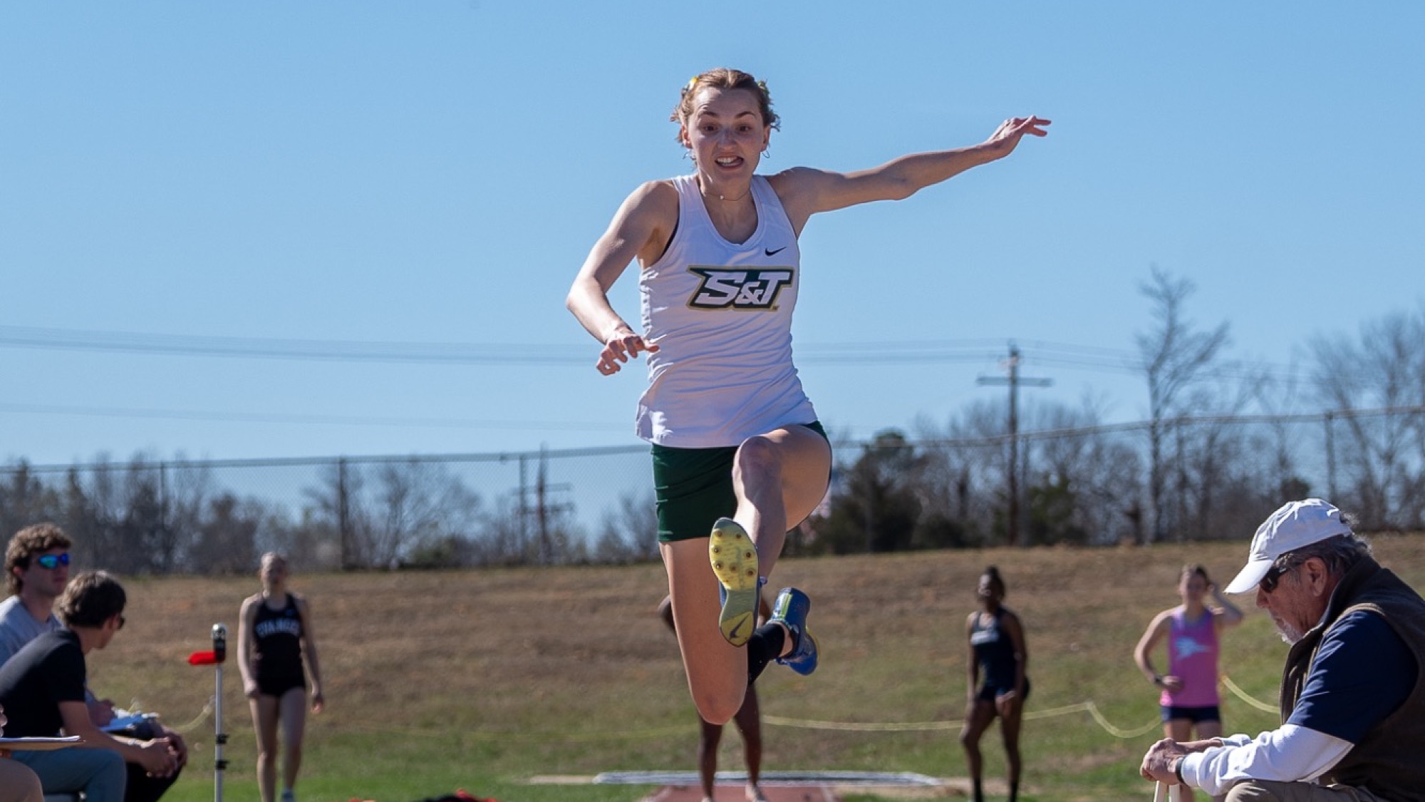 Missouri S&T track and field athlete Ali McClure soars through the air during the long jump at the Miner Invitational, approaching the sand pit as officials and competitors look on from the runway.