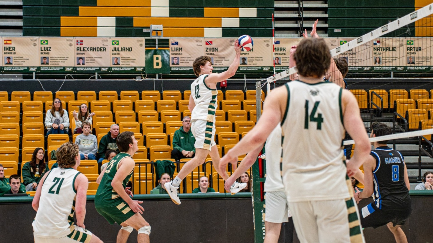 Camden Seaver, wearing a white Missouri S&T volleyball uniform with number 12, is photographed mid-air at the net as he swings his right arm to attack the ball during a match. Teammates in white are positioned around him in ready stances, while opposing players in dark uniforms prepare to defend on the other side. The action takes place inside a gymnasium with green and gold seating and championship signage visible in the background.