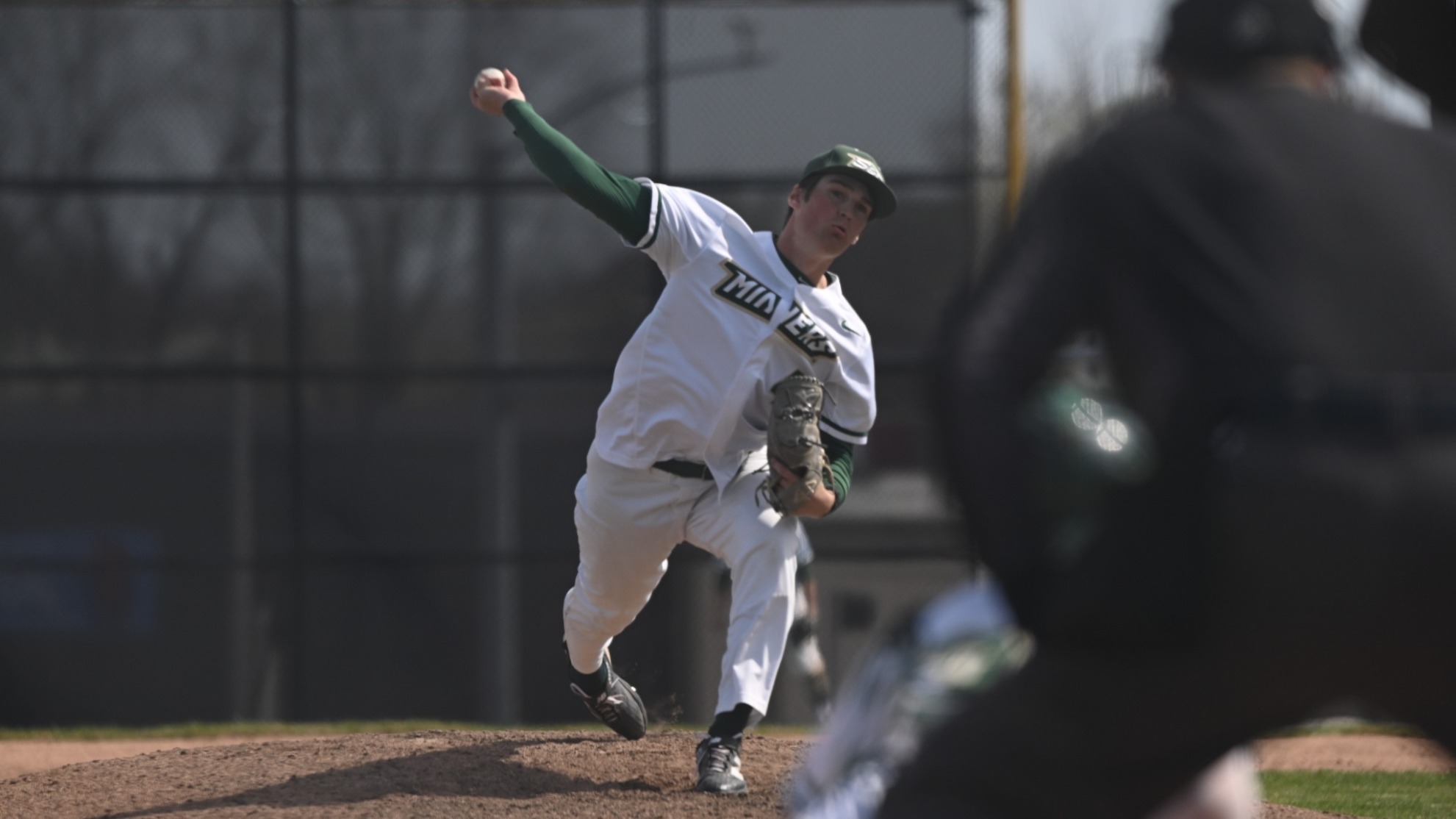 Jack Manninen, a Missouri S&T pitcher, is photographed in the middle of his pitching motion on a dirt mound, striding forward off his back leg with his throwing arm fully extended as he releases the baseball toward home plate. He wears a white Miners uniform with green sleeves and a green cap, his glove hand pulled in near his torso as his body leans toward the plate. In the foreground, a blurred opposing batter and catcher frame the action, while a dark outfield fence and trees sit in the background under daylight conditions.