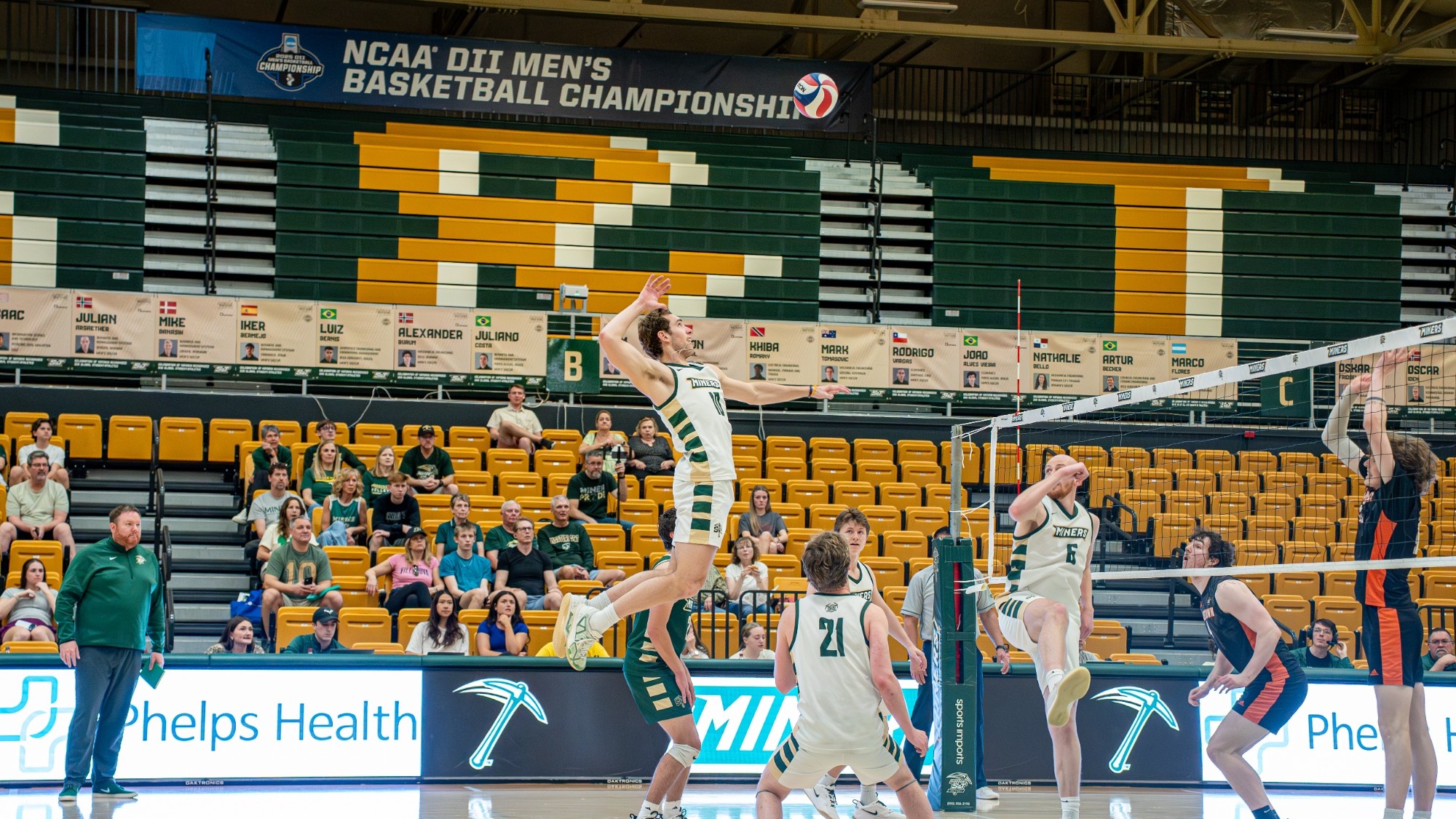 Missouri S&T men’s volleyball player Nate Meyer, wearing a white Miners uniform, is photographed leaping high above the net to spike the ball with his right hand during a match inside Gibson Arena. His body is fully extended in midair, with his hitting arm drawn back and eyes focused on the ball above him. Teammates in white uniforms are positioned around the court preparing for the play, including one near the net and others in defensive stances. Two opposing players in dark uniforms jump to block on the right side of the net.