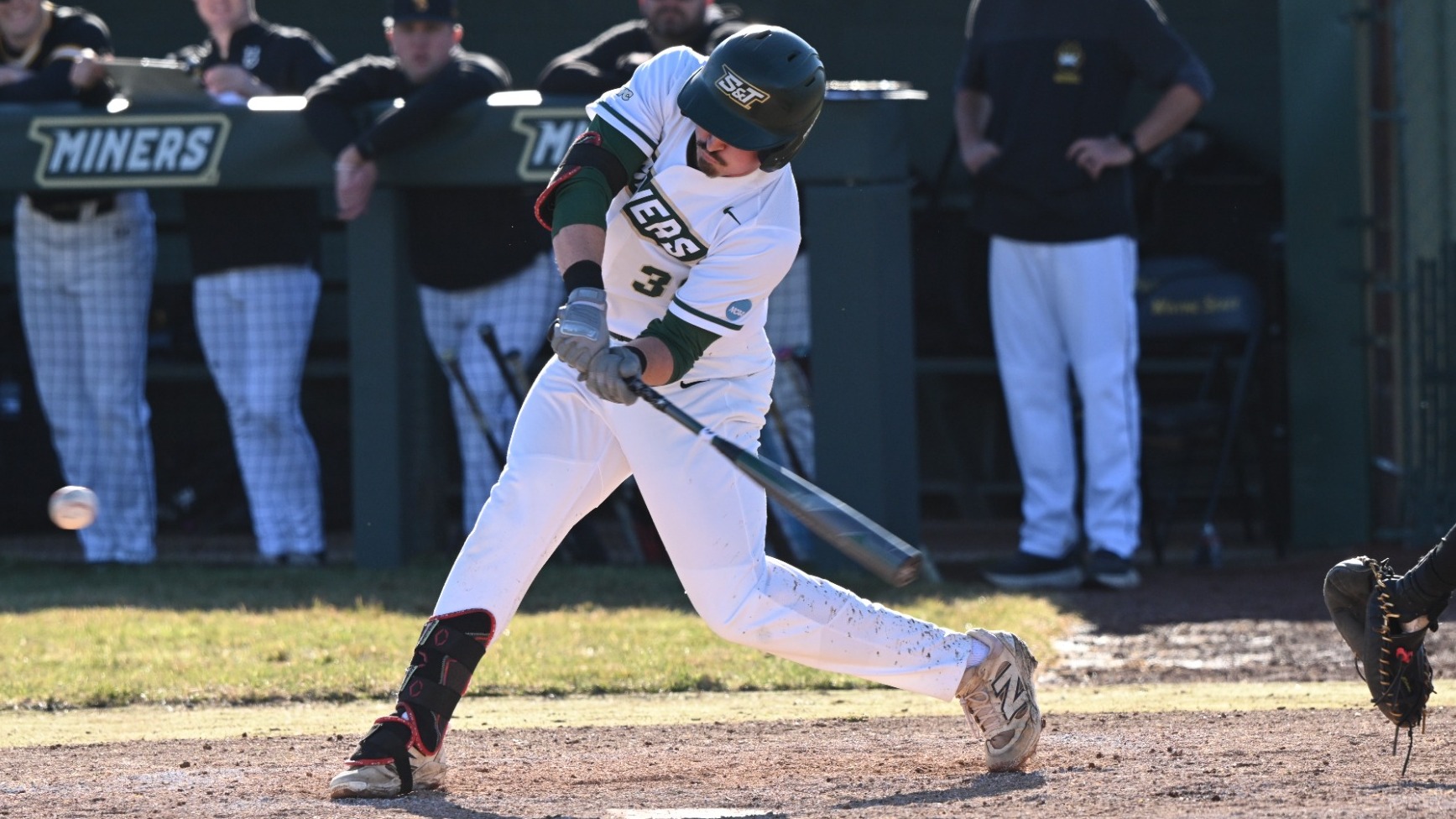 Missouri S&T baseball player Caleb Horsey, wearing a white Miners uniform with green lettering and number 3, is photographed making contact with the ball during a swing at home plate. The ball is just leaving the bat as he follows through, with dirt kicking up around his front foot. A catcher in dark gear is positioned to the right