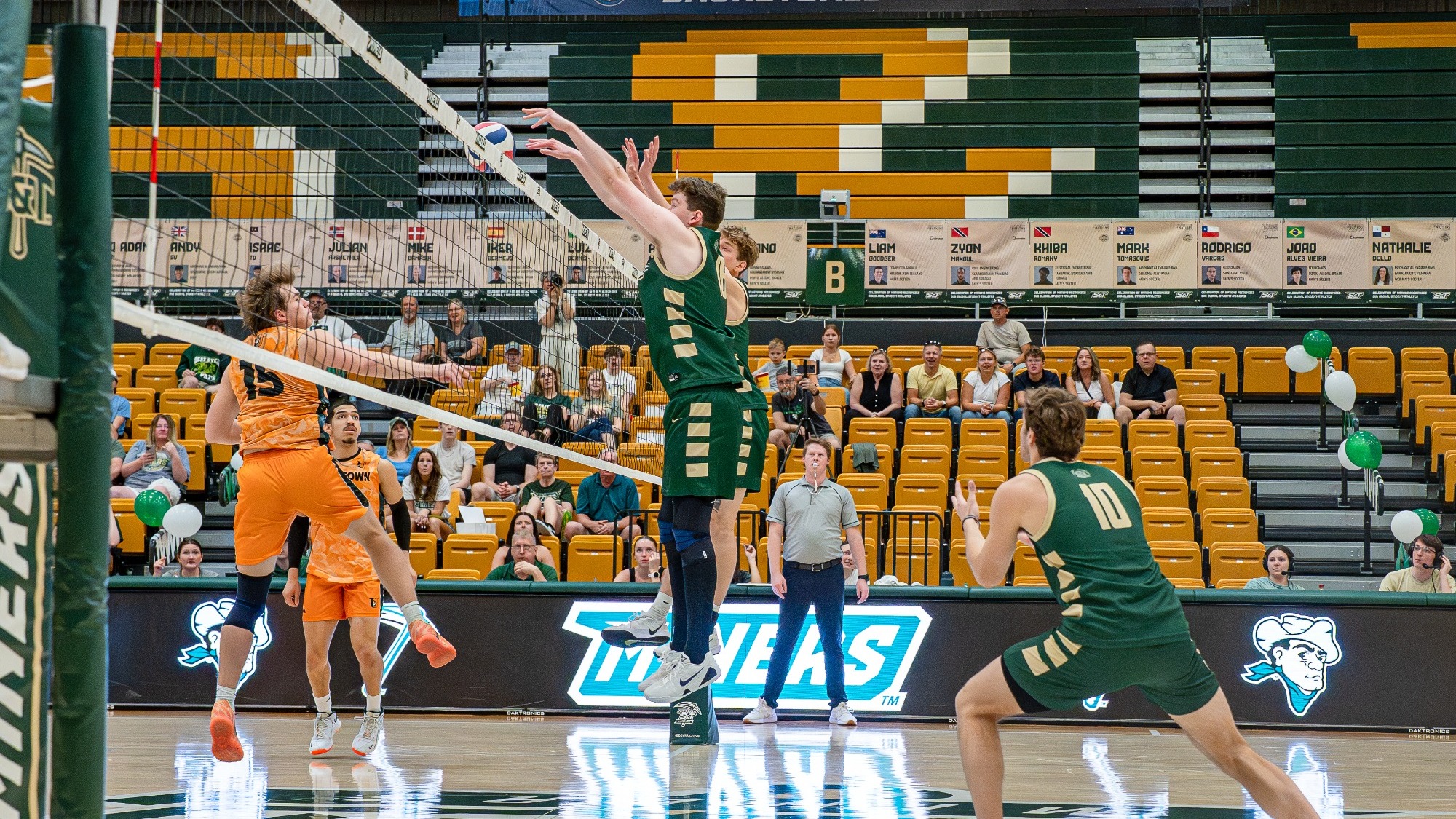 Grogan Kraus and Erik Giezycki, Missouri S&T men's volleyball players are wearing green uniforms and jumping at the net to block an attack by a jamestown opponent during a match in Gibson Arena