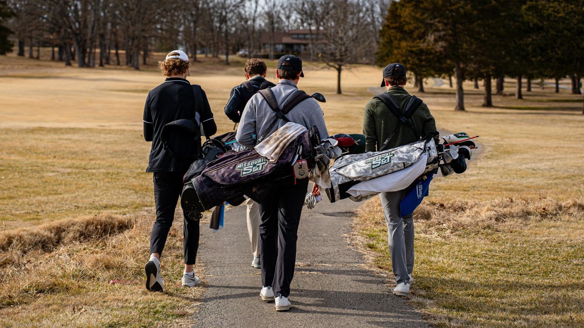 Four Missouri S&T golfers are photographed from behind walking down a paved cart path on a golf course, each carrying a golf bag over their shoulder. The group heads toward the fairway surrounded by dormant grass and leafless trees, with a clubhouse visible in the distance under a clear sky.