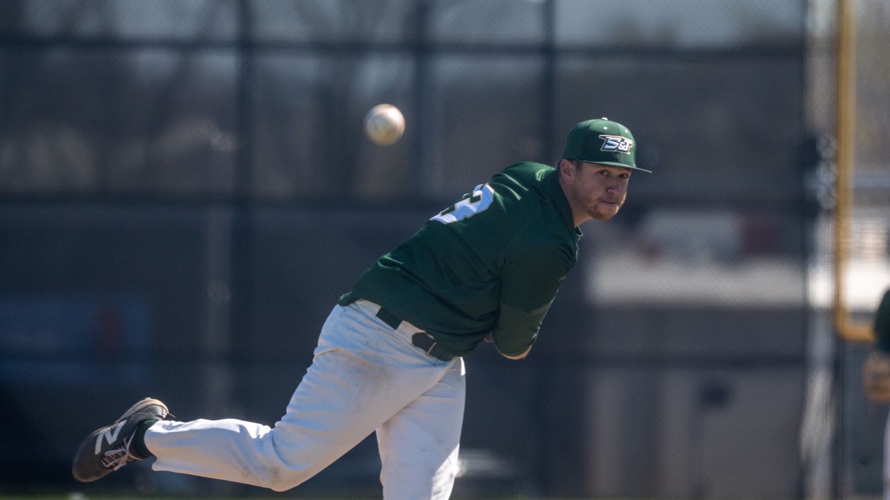 Dylan Bates a Missouri S&T pitcher is thrown mid follow through after throwing a baseball. The baseball is out of focus in the middle of the frame. Bates is wearing a green Miners baseball uniform with a green cap that has the S&T logo on it