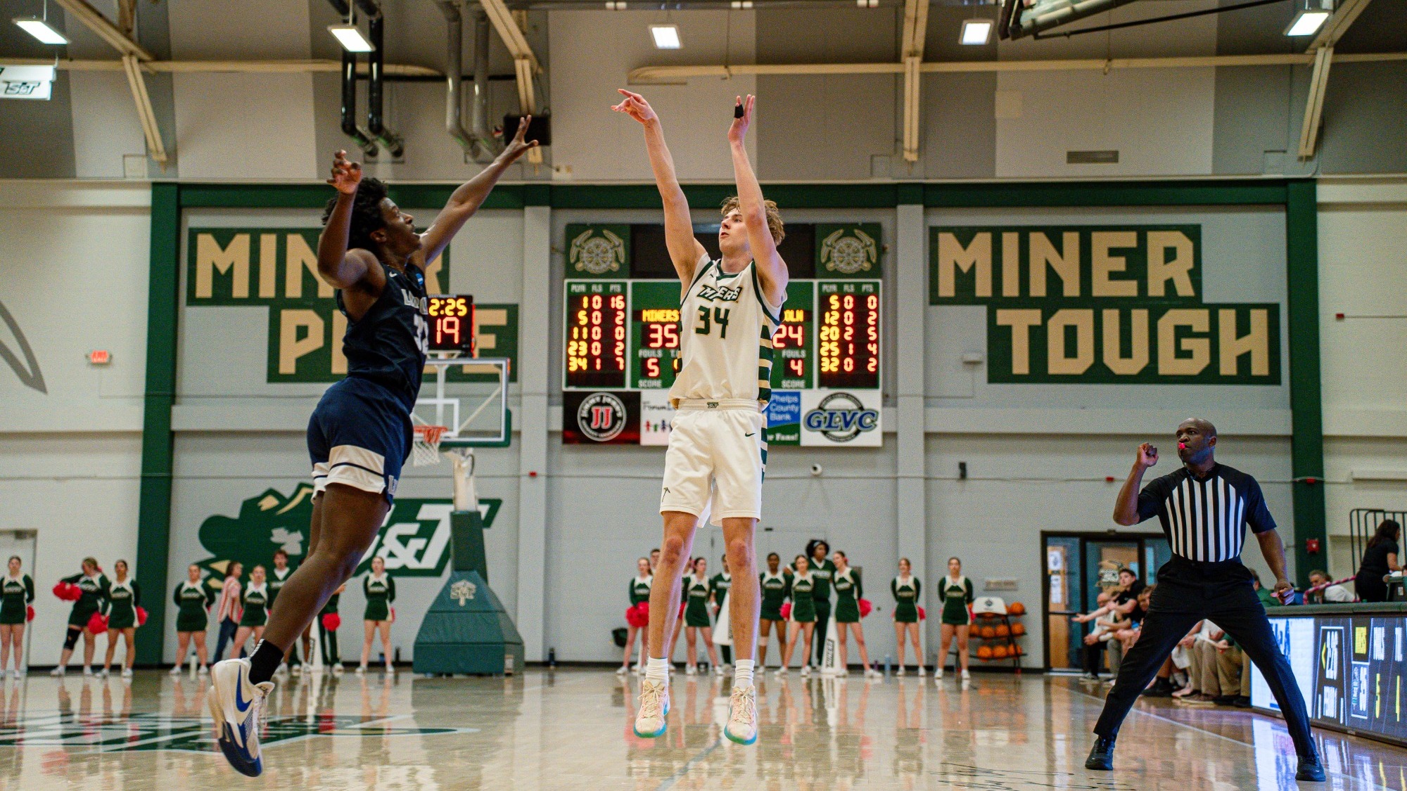 Andrew Young, wearing a white Missouri S&T uniform (#34), is photographed mid-air releasing a jump shot during a basketball game, with a defender in a dark uniform leaping to contest the shot. The ball is visible above his outstretched arms as it travels toward the basket. A referee stands to the right signaling the play, while cheerleaders and a scoreboard showing game time and score are visible in the background inside the gym.
