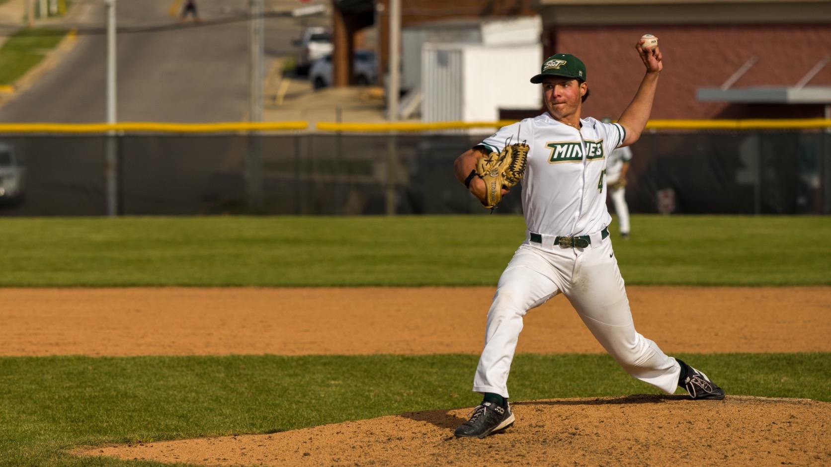 A baseball pitcher in a white “Miners” uniform, identified as Drew Timp, stands on the mound mid-throw with his right arm raised and glove hand forward. His body is extended in a pitching stride, focused toward home plate. The field’s grass and dirt mound are clearly visible, with an outfield fence, buildings, and a street in the background.