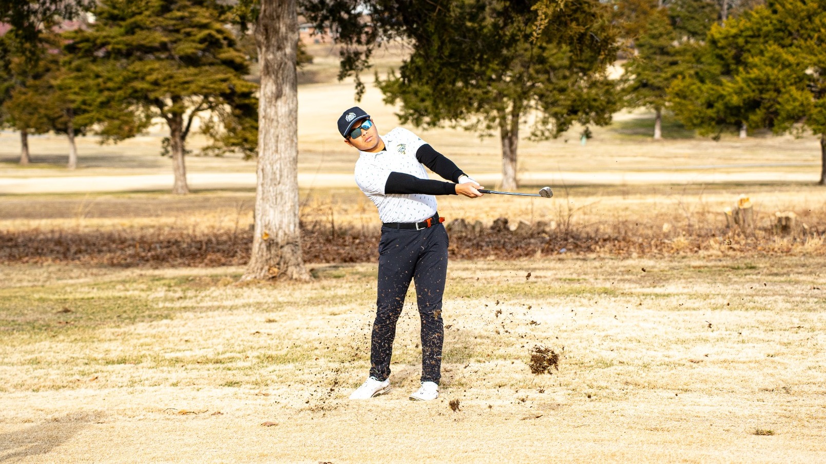 Golfer Rizq Adam Rohizam completes a powerful swing on a dry, dormant grass fairway. He is wearing a white patterned polo, black compression sleeves, dark trousers, and a navy baseball cap with sunglasses. Dirt and turf are captured mid-air around his feet from the impact of the club. His golf bag stands to the left in the foreground, with a backdrop of sparse trees and a clear winter landscape.